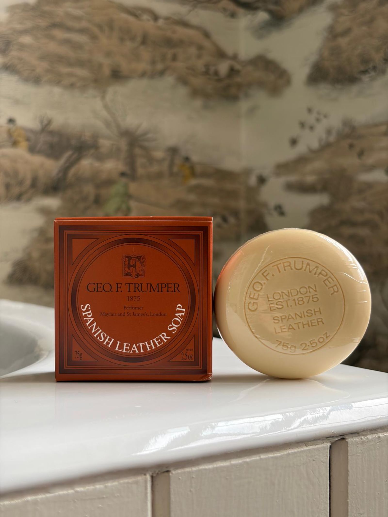 A set of 3 Geo. F. Trumper Spanish Leather Hand Soaps in their signature red box sits on white tiles, with a patterned wall depicting pastoral scenes in the background.