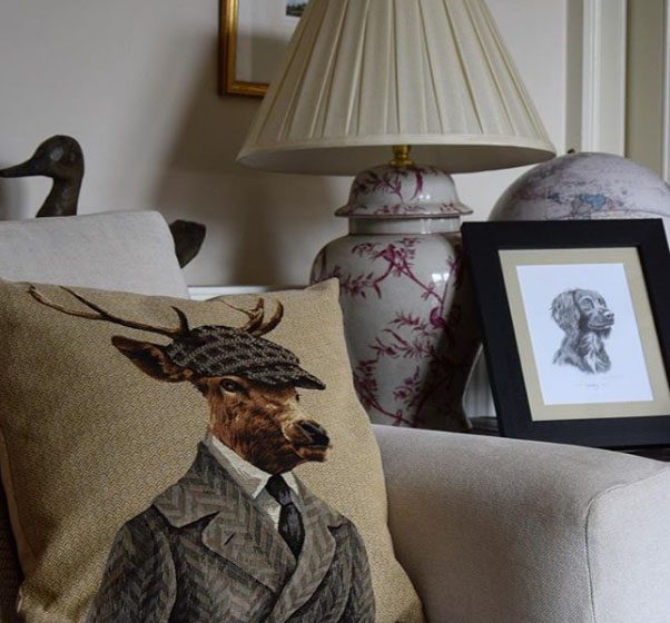 A beige sofa is adorned with Campbells of Beauly Dapper Stag Cushion Cover. Behind it, a lamp with a patterned base, framed dog sketch, globe, and wooden duck sculpture enhance the stylish home decor.