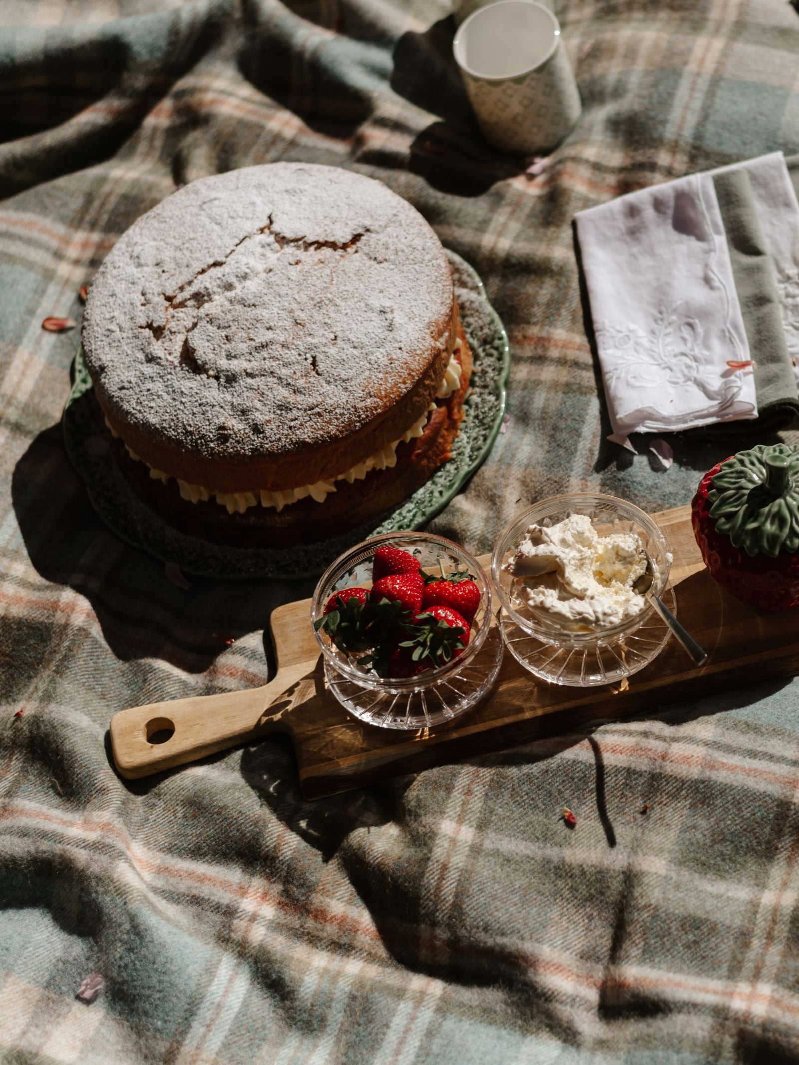 A sponge cake dusted with powdered sugar sits on a plate atop the Strathcarron Lambswool Throw by Campbell's of Beauly, next to a wooden board with bowls of strawberries and whipped cream, plus a cup and napkin nearby.