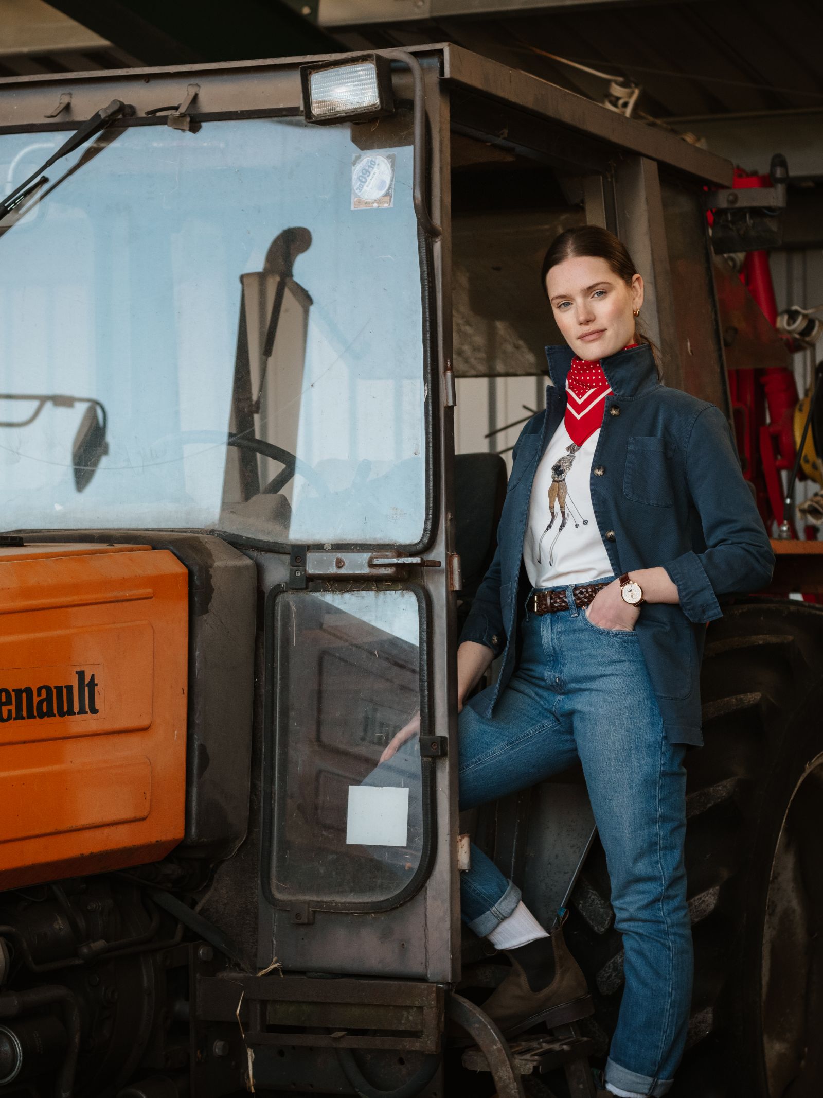 A woman in relaxed fit jeans, a Campbell's of Beauly Summer Chore Jacket, red scarf, and brown boots stands confidently by the open door of an orange Renault tractor inside a barn.