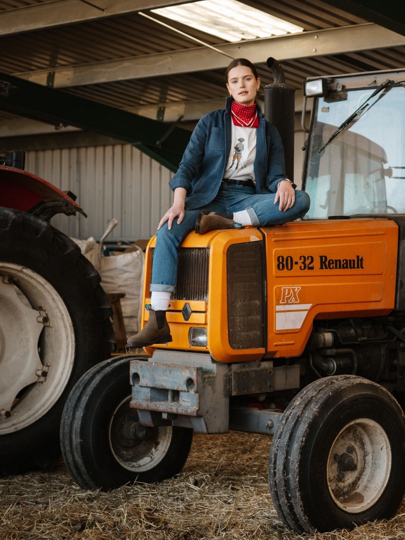 Wearing jeans, brown boots, a red bandana, and a Campbell's of Beauly Summer Chore Jacket, a person sits on the hood of an orange Renault 80-32 PX tractor inside a barn with another tractor in the background.
