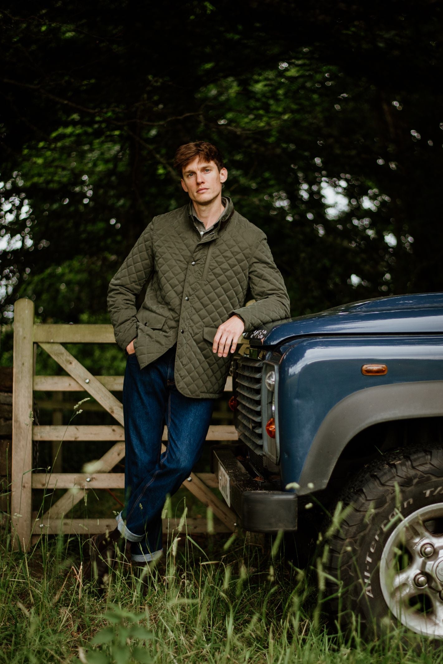 Wearing the Campbell's of Beauly Auld Stock Tomich Jacket and blue jeans, a person leans against a blue off-road vehicle near a wooden gate amid tall grass and trees—capturing classic country style.
