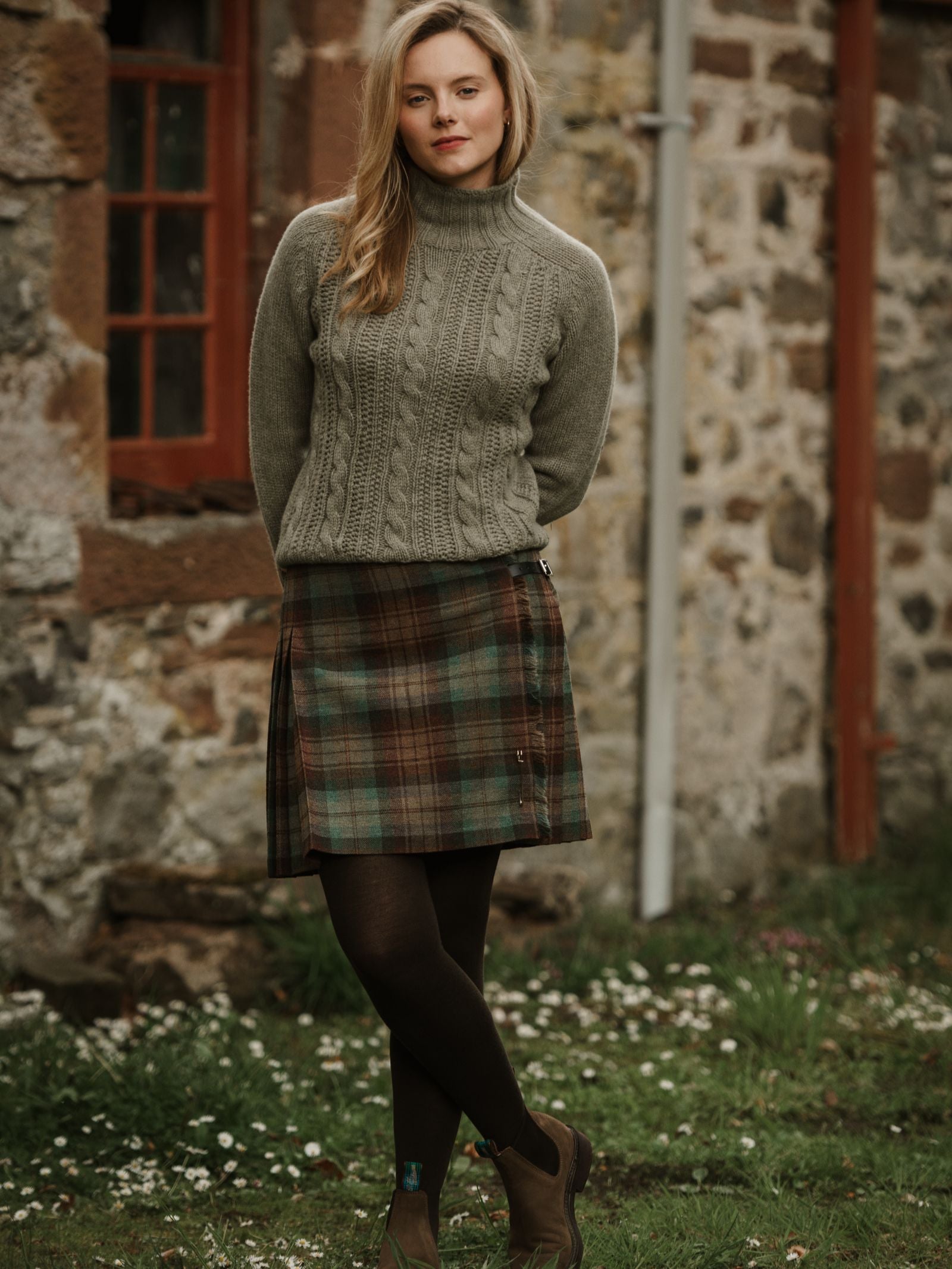 A woman with blonde hair poses outdoors on grass in front of a stone building, wearing the Campbell's of Beauly Tweed Kilt, a gray cable-knit sweater, dark tights, and brown boots.