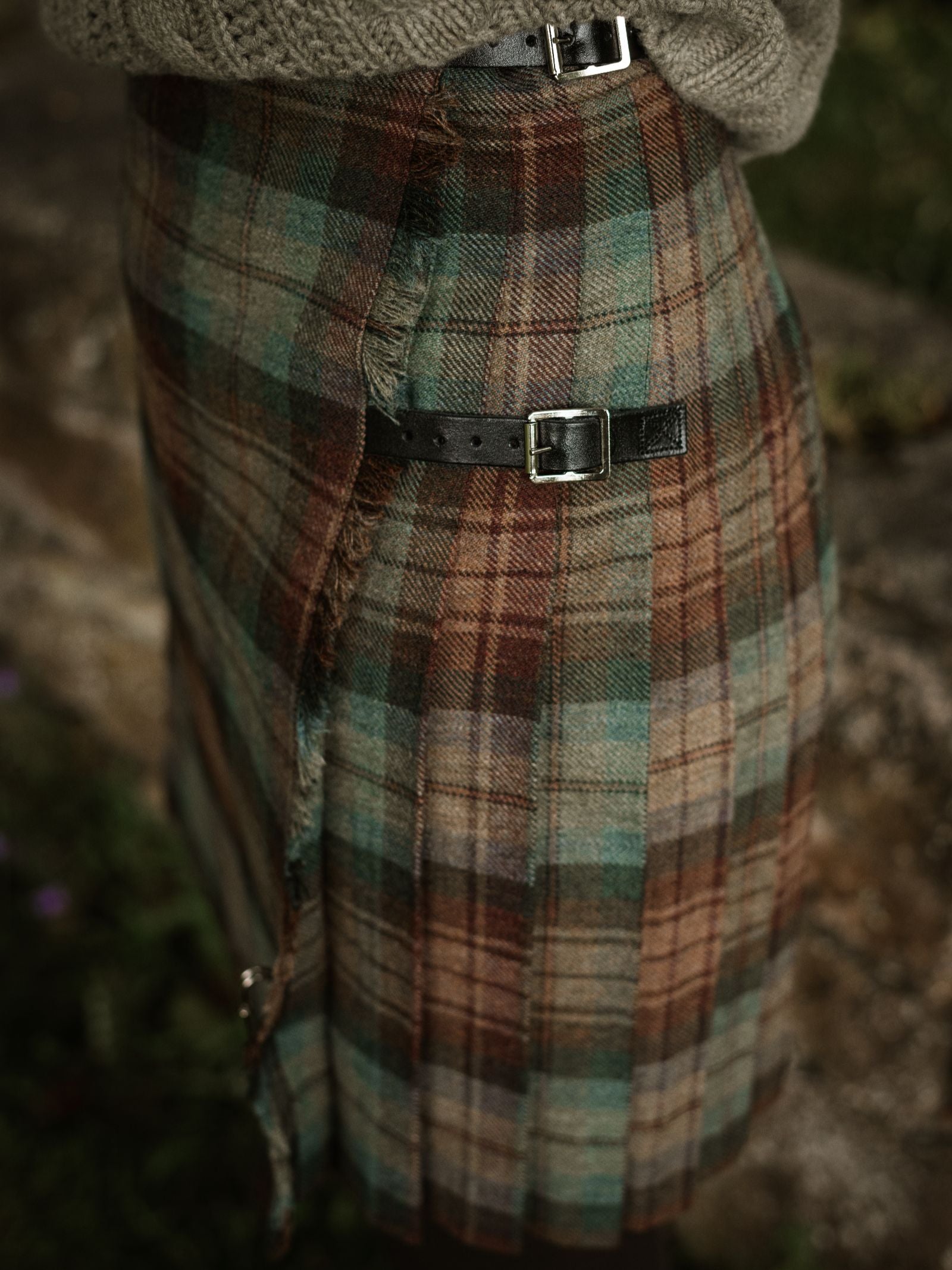 A close-up shows a person wearing the Campbell's of Beauly Tweed Kilt in green, brown, and beige tartan, featuring two black leather buckles and paired with a grey knitted sweater.