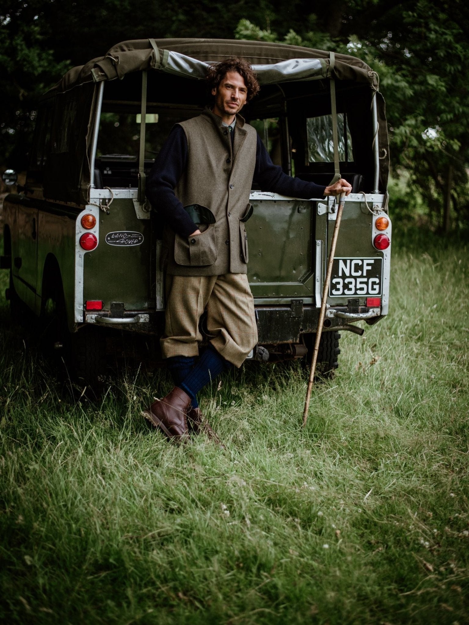 Leaning against an old green Land Rover in a grassy field, a person in country attire wears Campbell’s of Beauly Plus Fours, holding a walking stick and cap among the trees for a classic vintage outdoor look.