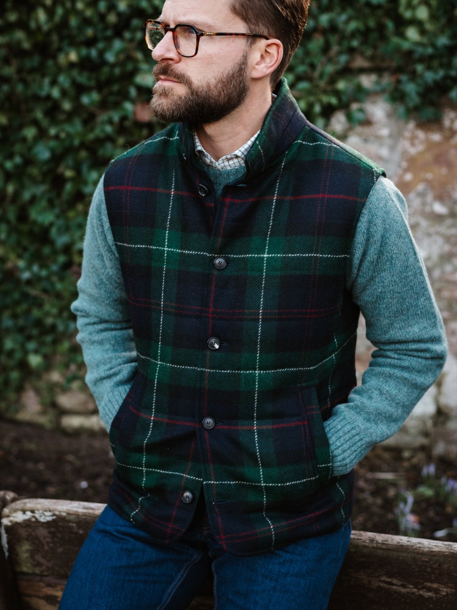 A man with a beard and glasses wears the Campbell's of Beauly Auld Stock Quilted Tweed Buttoned Gilet over a blue-gray sweater and jeans, sitting outdoors on a bench with an ivy-covered wall, showcasing the perfect layering piece.