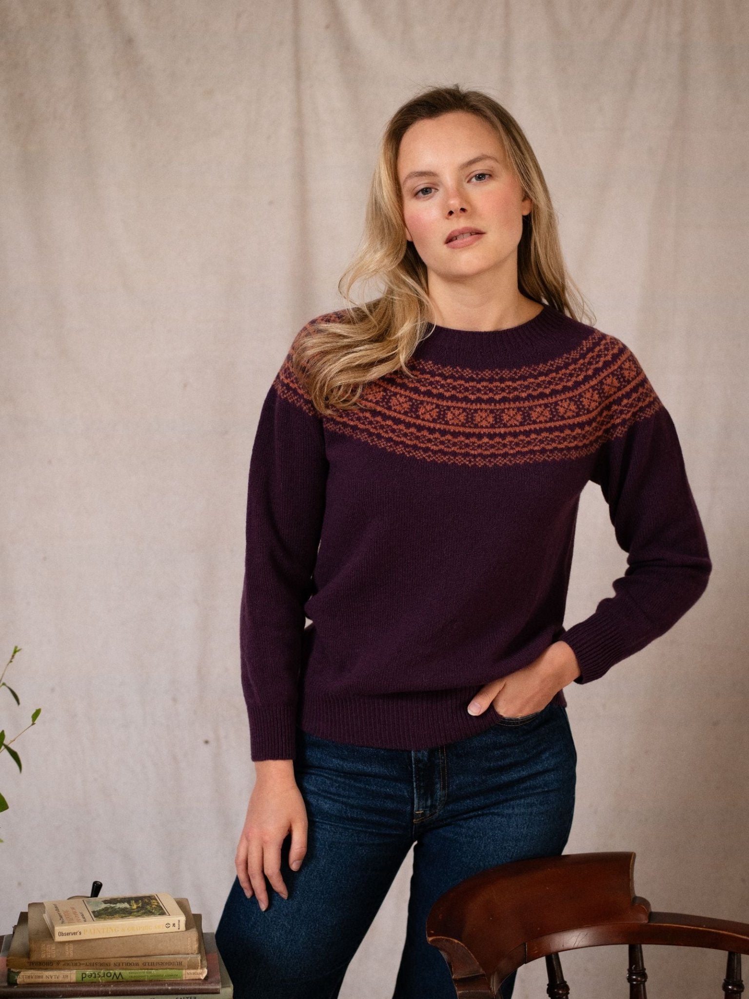 A woman with long blonde hair wears the Boysenberry Two-Colour Fairisle Yoke Jumper by Campbell's of Beauly and blue jeans, standing indoors beside a wooden chair and books, against a light fabric backdrop.