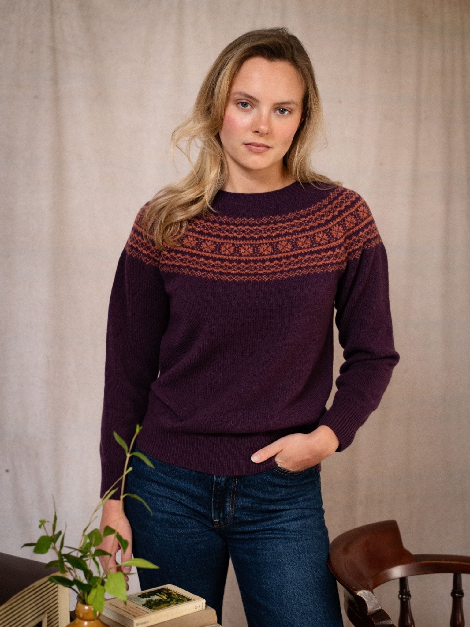 A woman with blonde hair wears the Campbell's of Beauly Two-Colour Fairisle Yoke Jumper in soft Geelong lambswool, standing indoors by a table with books and a potted plant, looking directly at the camera with a neutral expression.
