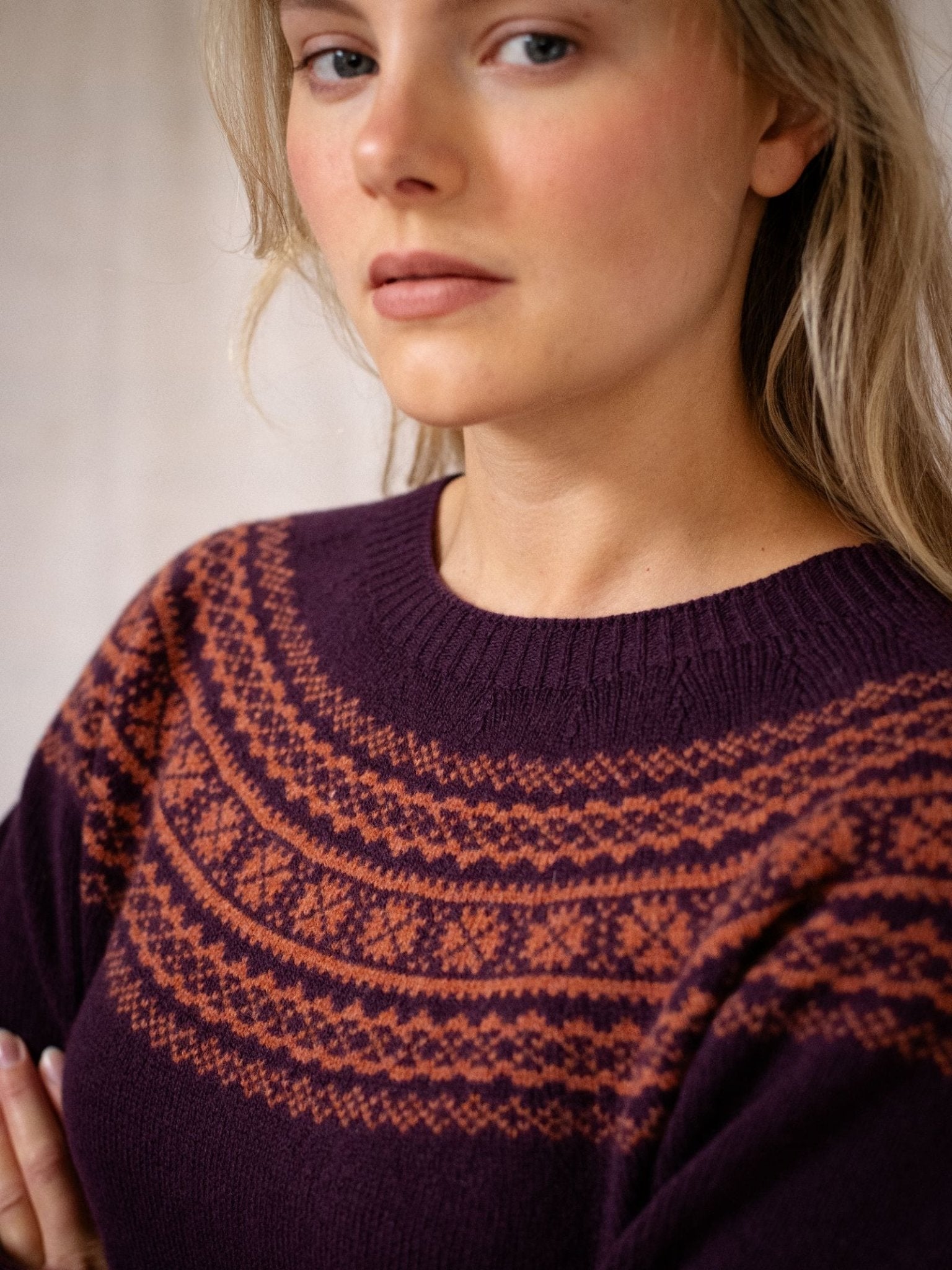 A woman with light skin and blonde hair wears the Campbell's of Beauly Two-Colour Fairisle Yoke Jumper, featuring a dark purple base and orange Scottish-inspired geometric pattern. She gazes slightly to the side with a neutral expression.