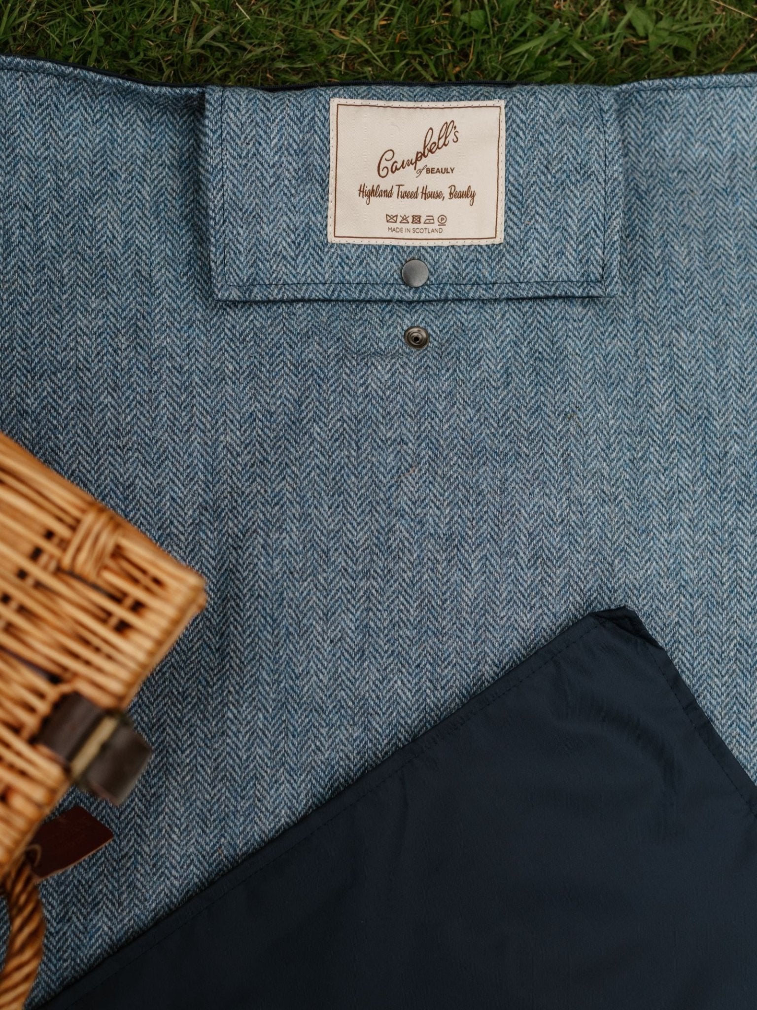 A close-up of the Walker Camper Picnic Rug by Campbell's of Beauly, featuring blue herringbone-patterned British wool and a branded label, shown beside a wicker basket and navy cloth on grass.