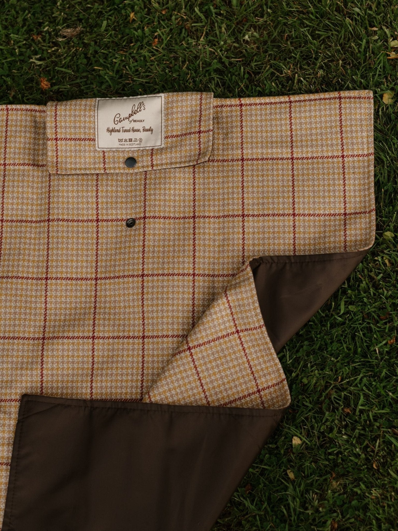 The Walker Camper Picnic Rug by Campbell's of Beauly, featuring a red and beige checkered pattern with brown lining, is laid flat on green grass. The brand label is partially visible at the edge.