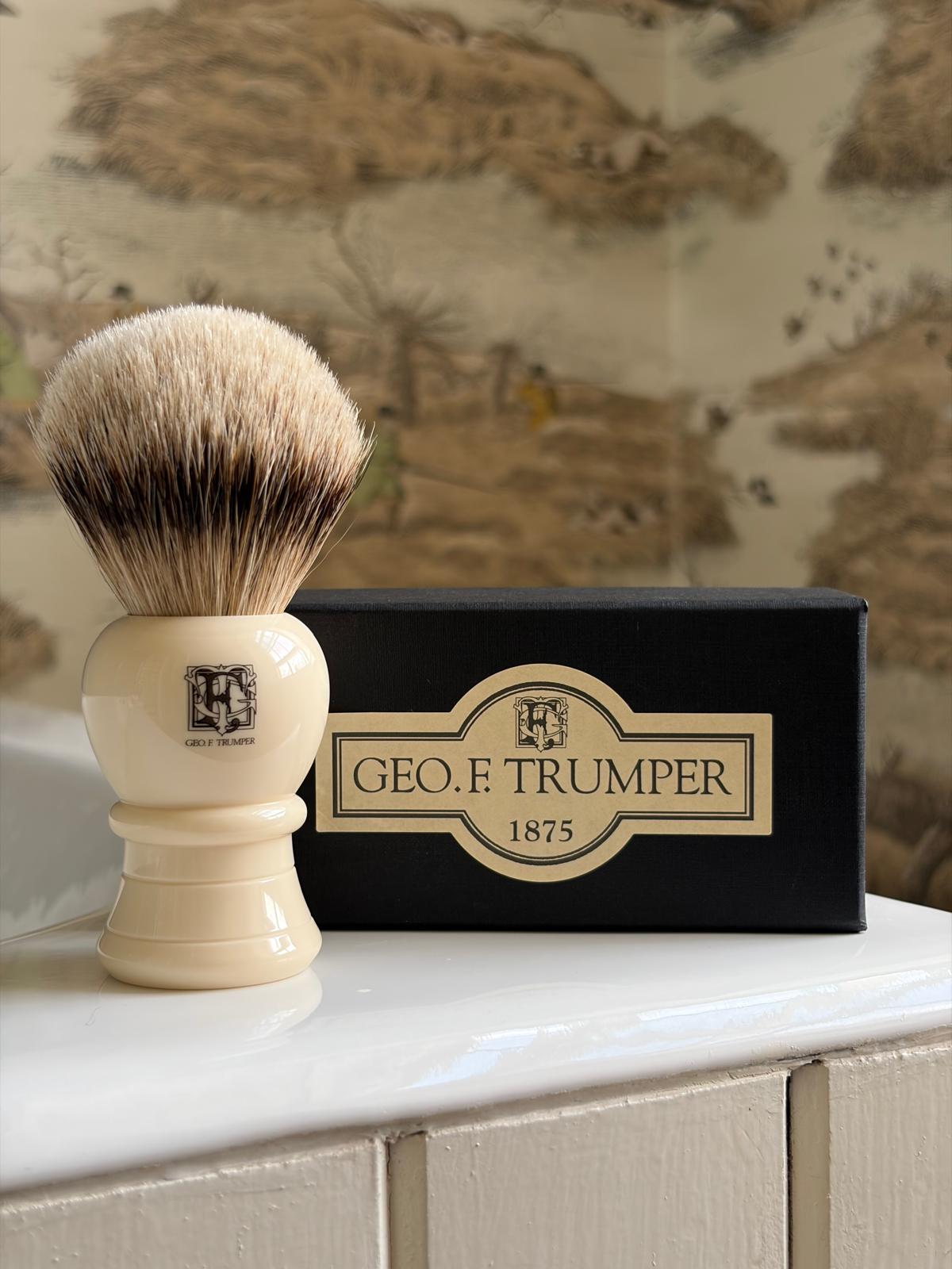 A Geo. F. Trumper Shaving Brush with a cream handle stands beside a black 1875 box on white tiles, offering a rich lather and classic wet shave; patterned wallpaper completes the scene.