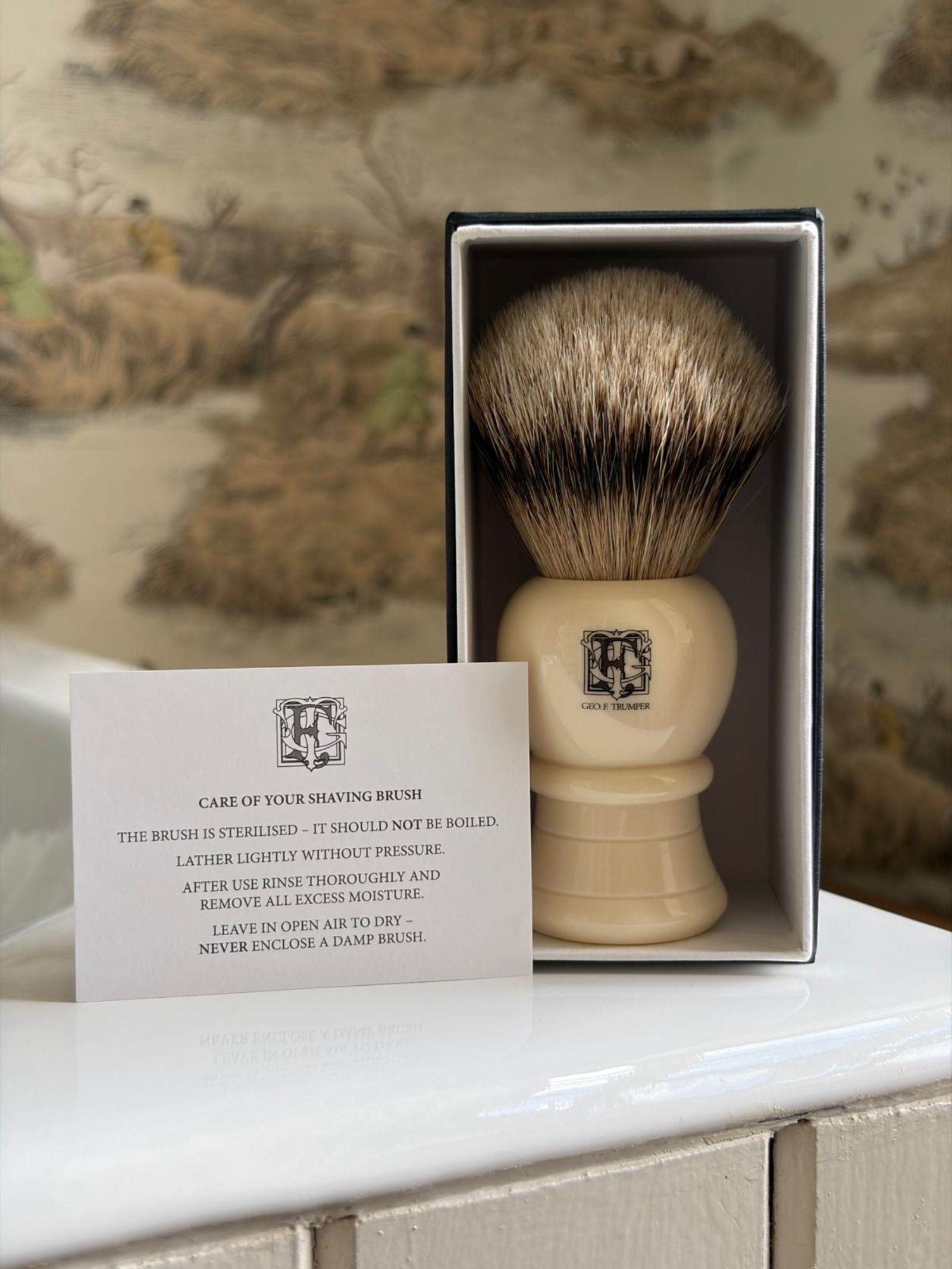 The Geo. F. Trumper Shaving Brush with a cream handle stands upright in its open box, accompanied by a care instruction card on cleaning and drying, set against a decorative background for a classic wet shave experience.