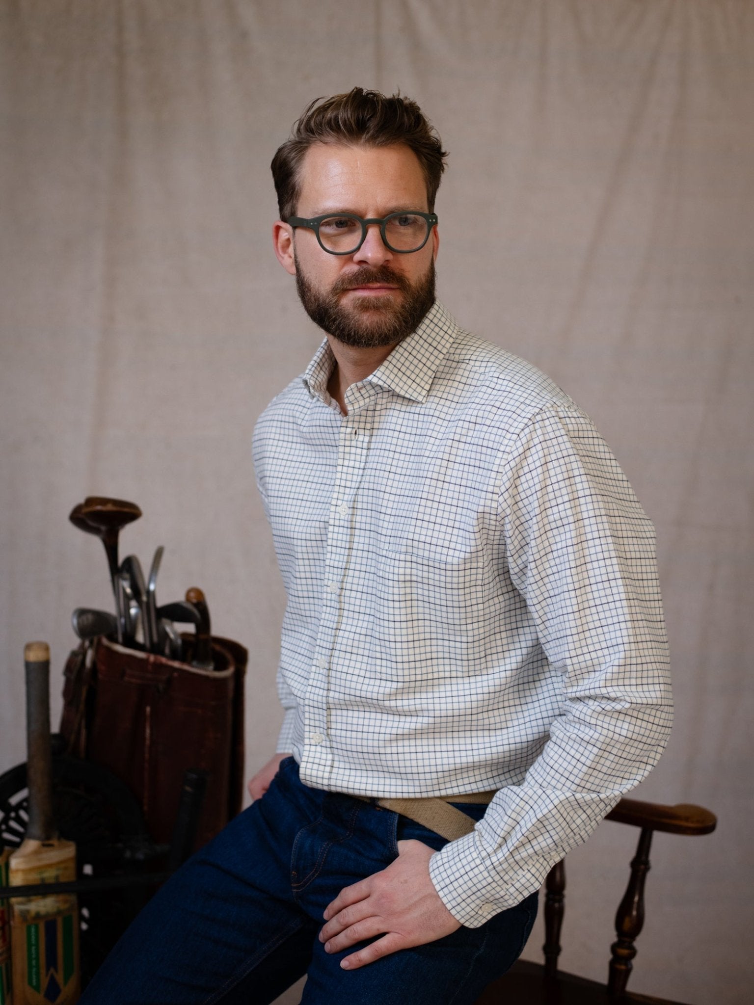 A bearded man in glasses wears the Campbell's of Beauly Windowpane Shirt and sits on a wooden chair, one hand in his pocket. Behind him, a vintage golf bag with clubs and a bat leans against a neutral backdrop.