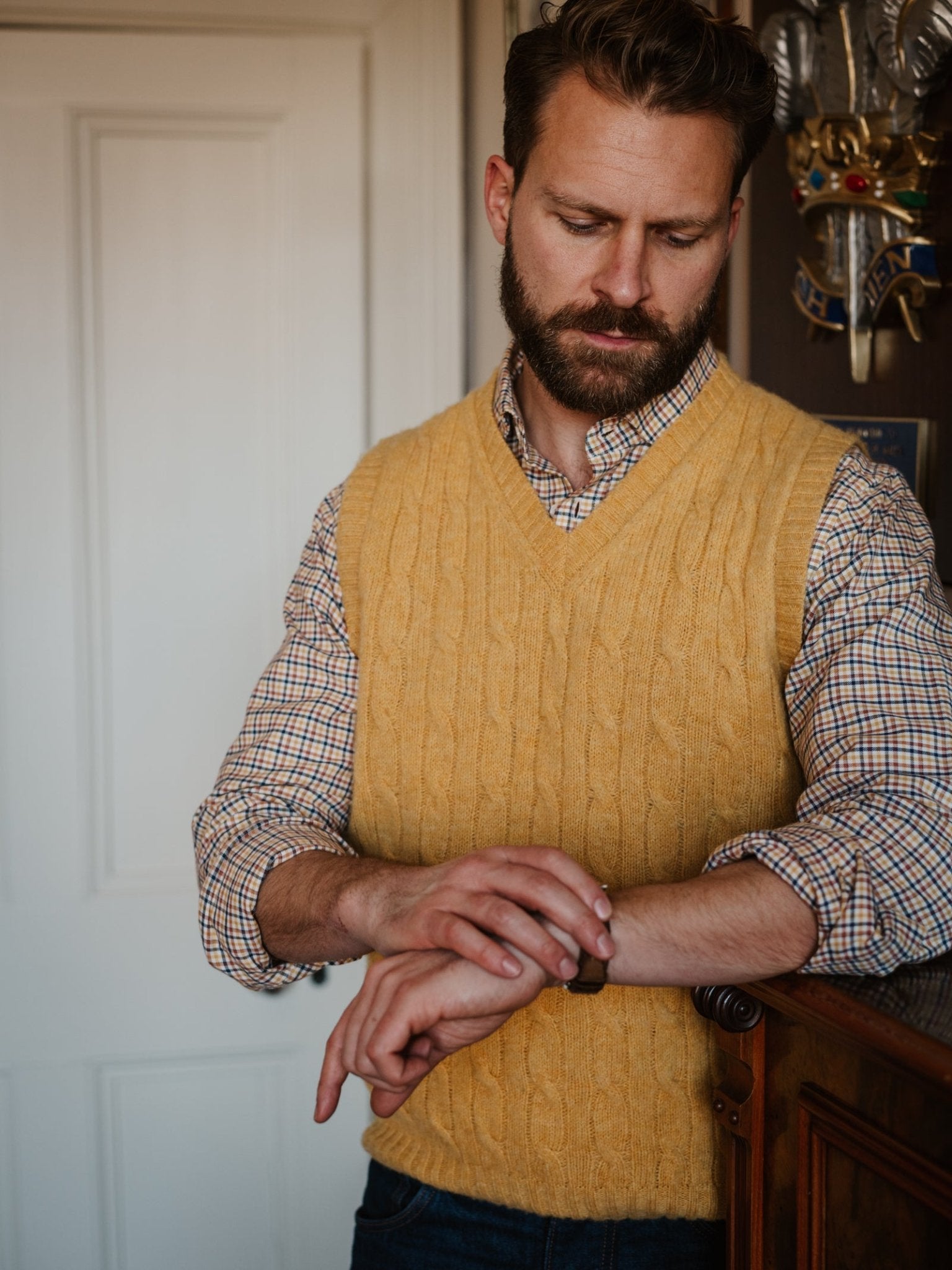 A bearded man in a Campbell’s of Beauly Windowpane Shirt wears a yellow cable-knit sweater vest and checks his watch, standing indoors near a white door and wooden surface.