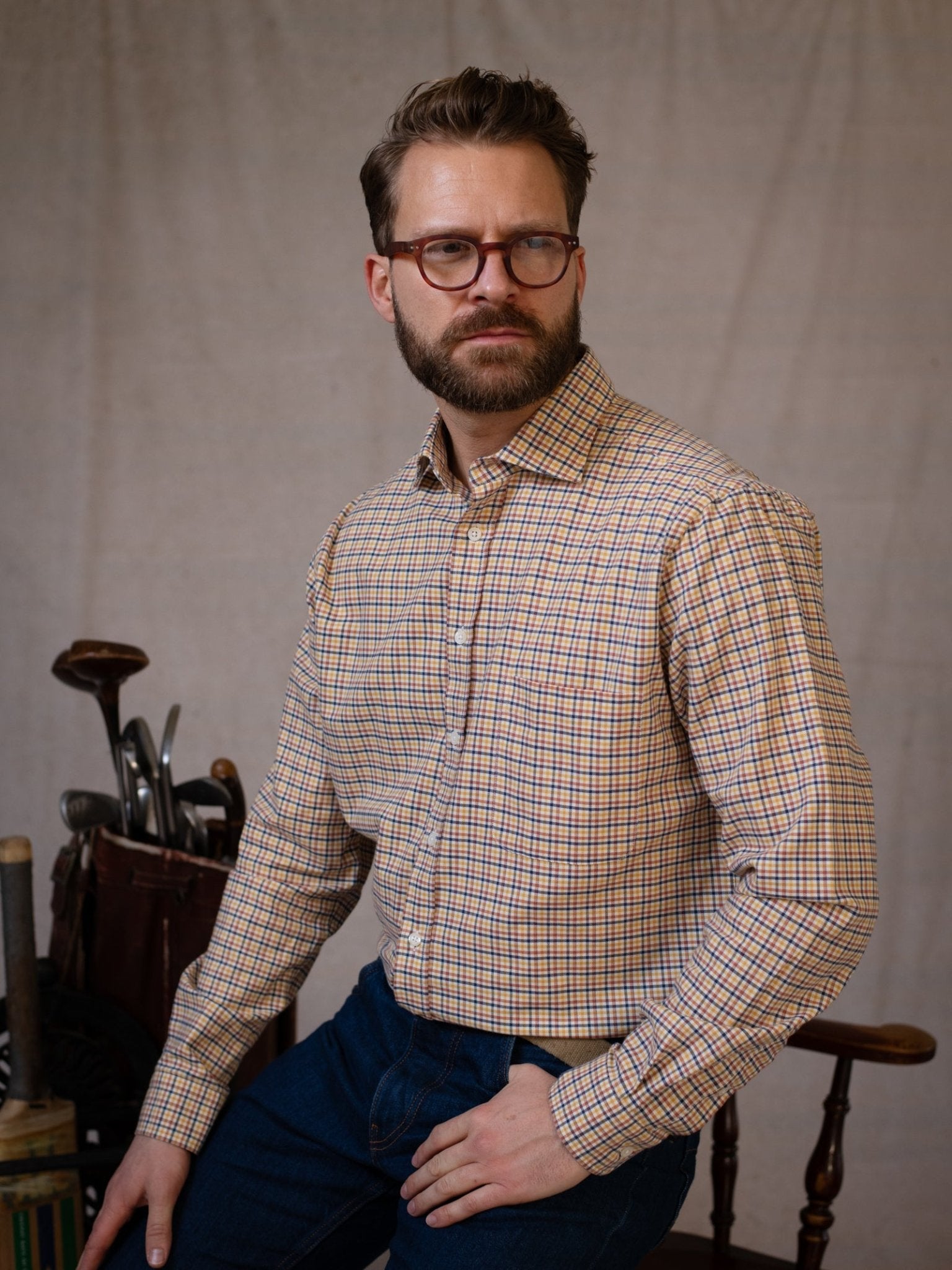 A bearded man with glasses, wearing the Campbell's of Beauly Windowpane Shirt and jeans, sits on a chair against a neutral background; a golf bag and clubs are in the corner.