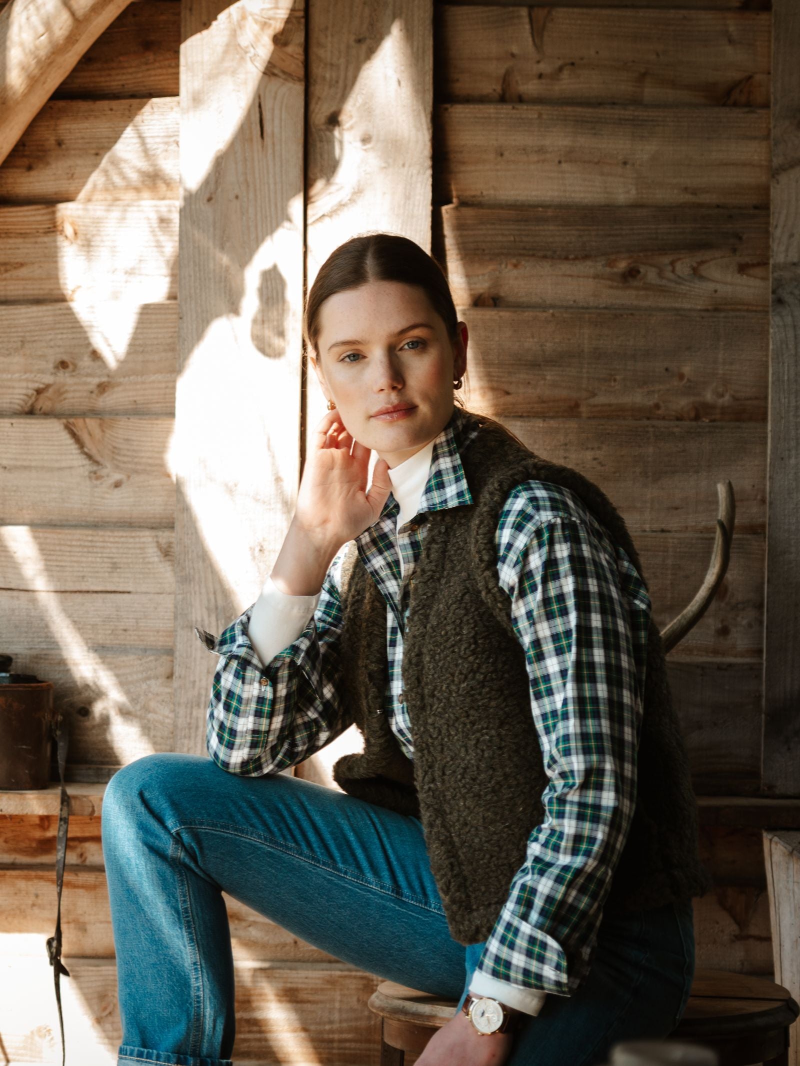 A woman with brown hair sits indoors by a wooden wall, wearing a plaid shirt, dark vest, and jeans. She rests her chin on her hand and gazes at the camera, illuminated by warm sunlight.