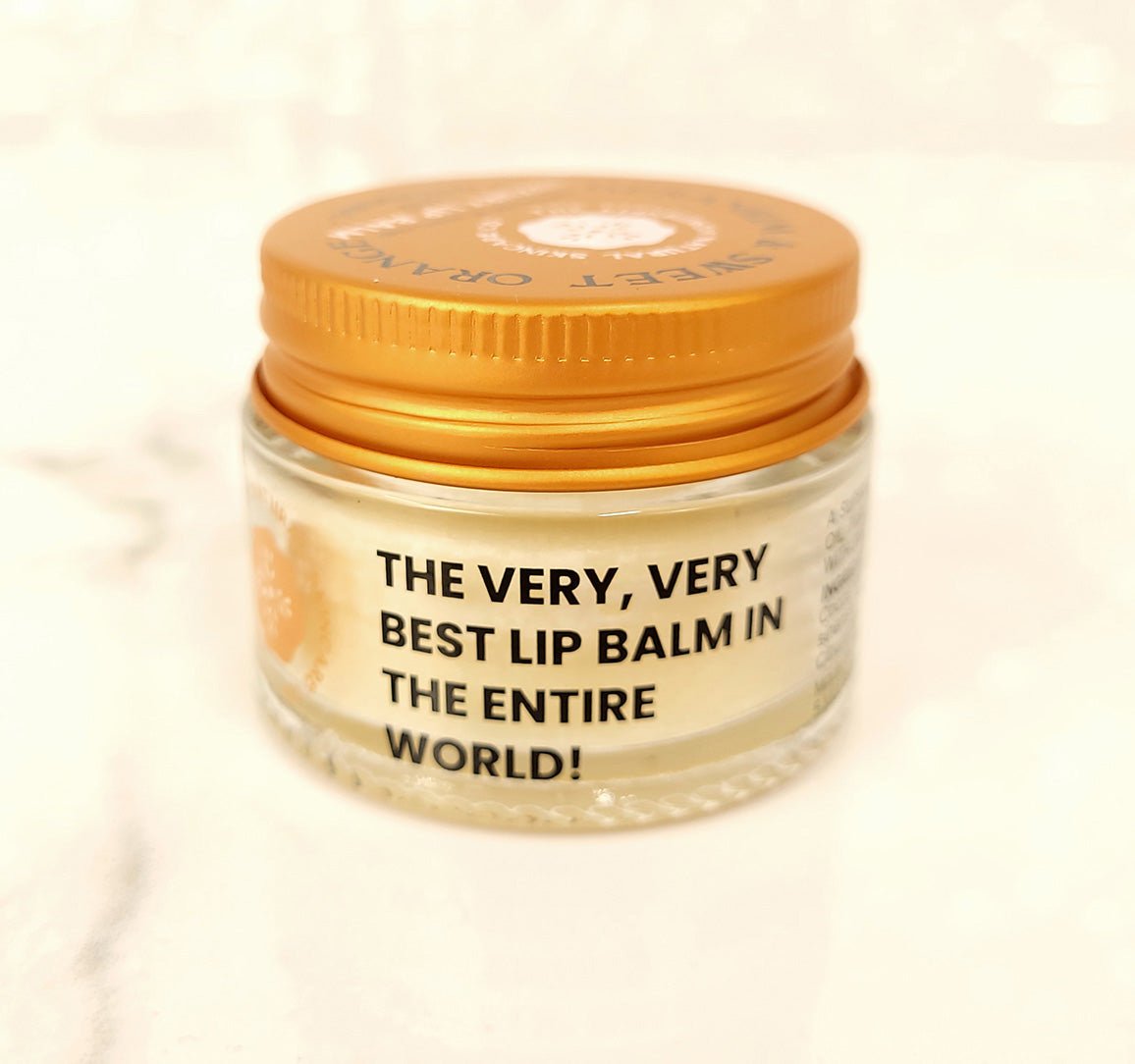 A small glass jar with an orange lid labeled THE VERY, VERY BEST LIP BALM IN THE ENTIRE WORLD! sits on a white surface. Its the Geranium & Sweet Orange Luxury Lip Balm by The Edinburgh Natural Skincare Company—100% natural, creamy, and boldly branded.