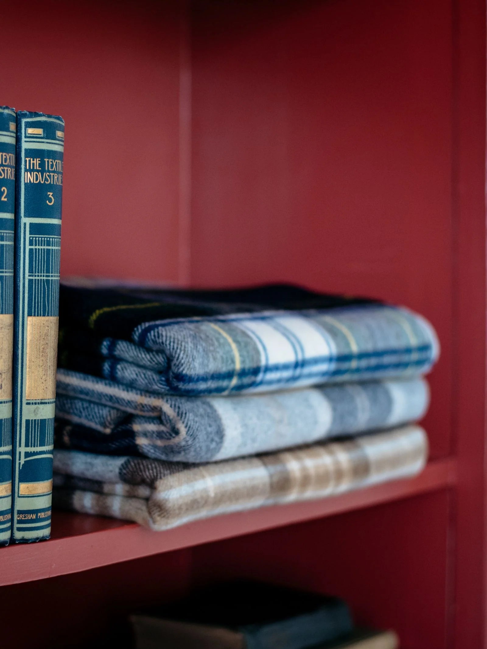 Three neatly folded Campbell's of Beauly Lambswool Tartan Blankets sit on a red shelf beside vintage blue books titled The Textile Industry, highlighting classic design and UK craftsmanship.