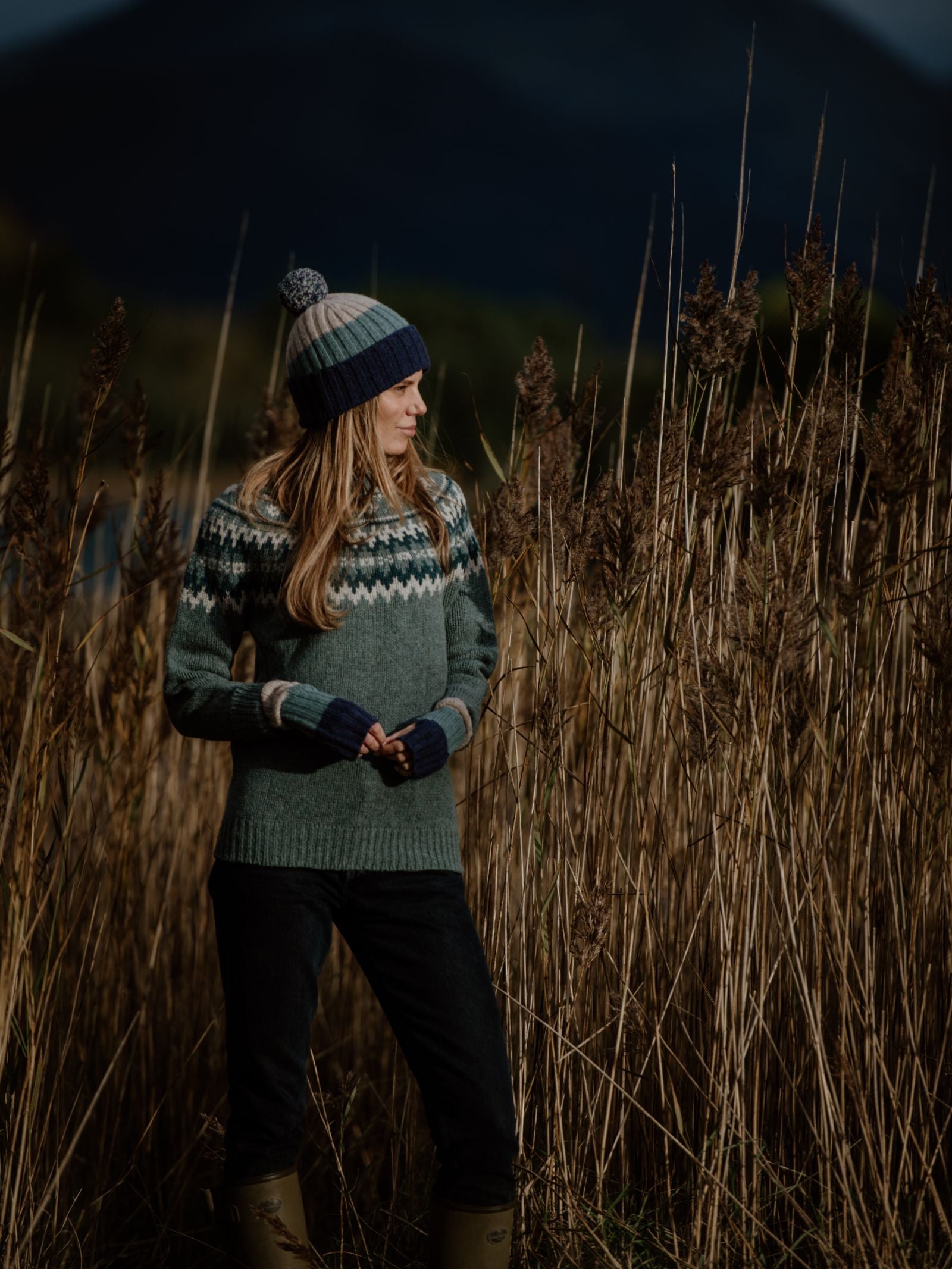 Wearing Auld Stock Stripe Wristwarmers by Campbell's of Beauly, a woman stands in tall dry grass amid dark mountains and clouds in the Scottish Borders, evoking an autumn or winter atmosphere.