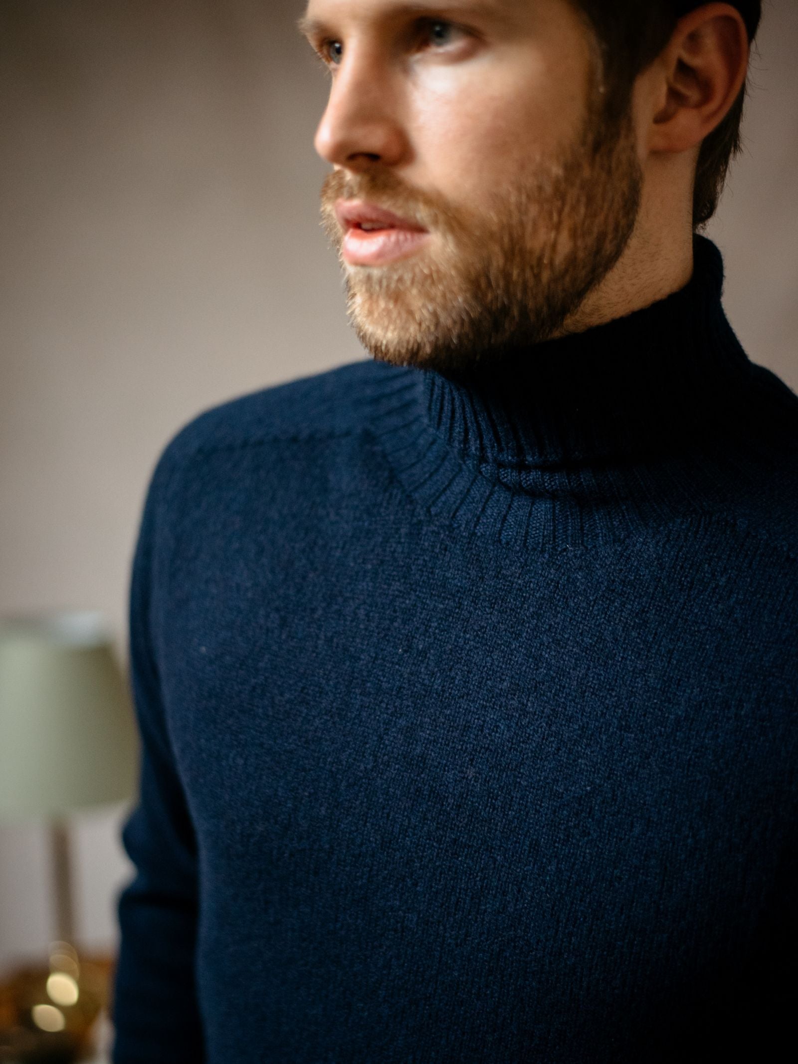A bearded man stands indoors wearing the Campbell's of Beauly Lambswool Roll Neck in dark blue. The close-up captures his upper body and face in partial profile with a blurred lamp in the background—ideal for layering.