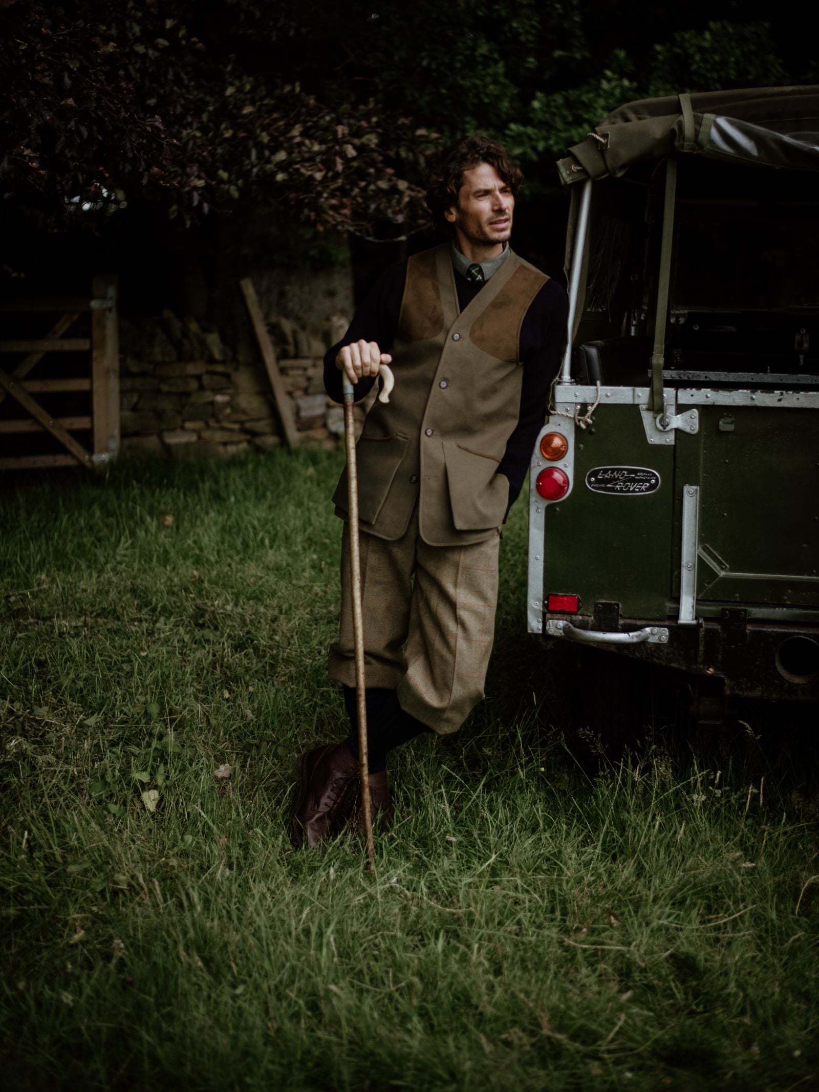 A man in country tweed and a Campbell's of Beauly Cotton Shooting Waistcoat leans on a walking stick by a green Land Rover, standing on grass near a wooden gate and stone wall, gazing thoughtfully into the distance.