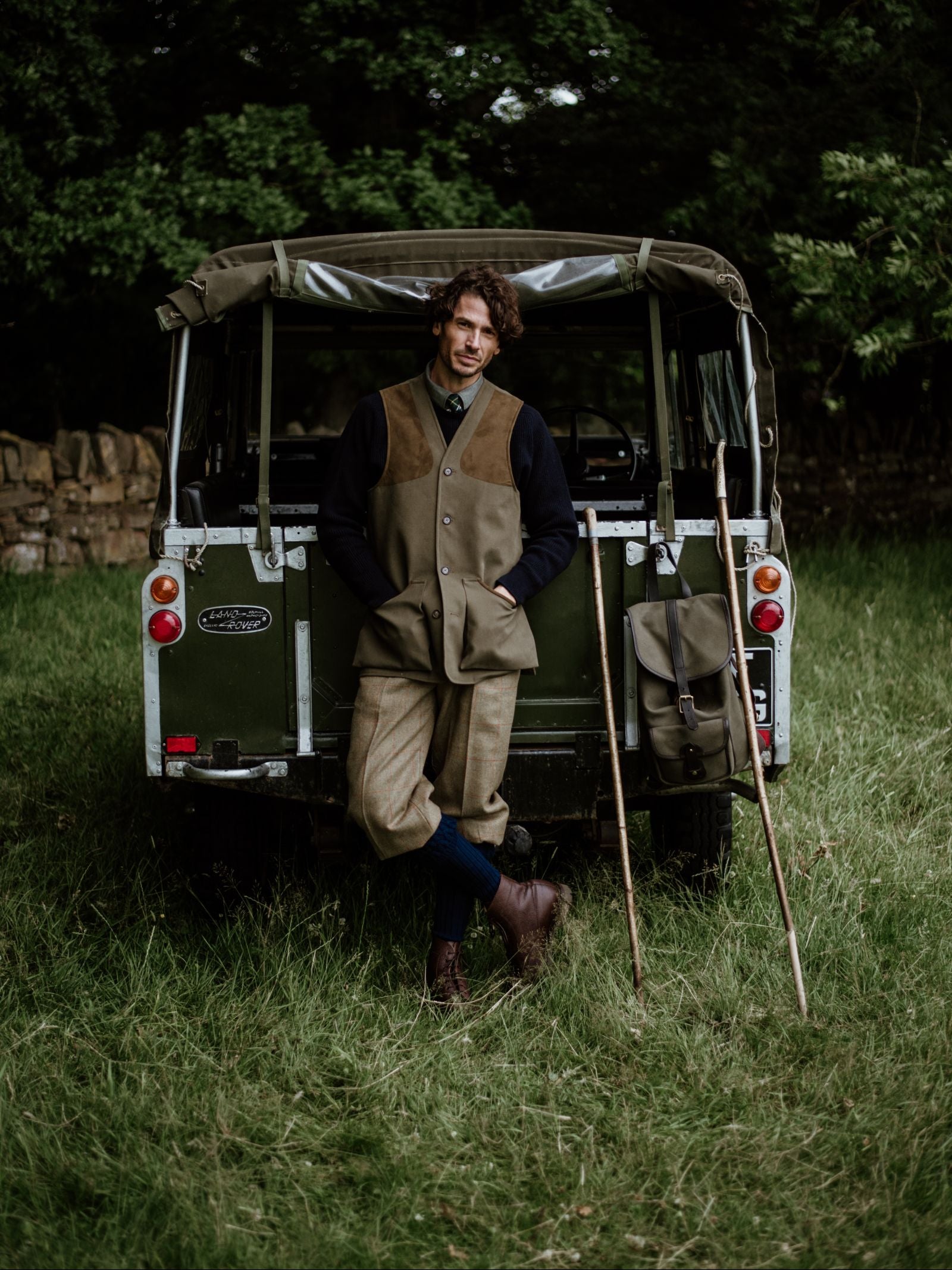 A man in country attire, wearing a Campbell's of Beauly Cotton Shooting Waistcoat, leans against a vintage green Land Rover on grass, with walking sticks and a satchel—ideal for countryside pursuits amid trees and a stone wall.