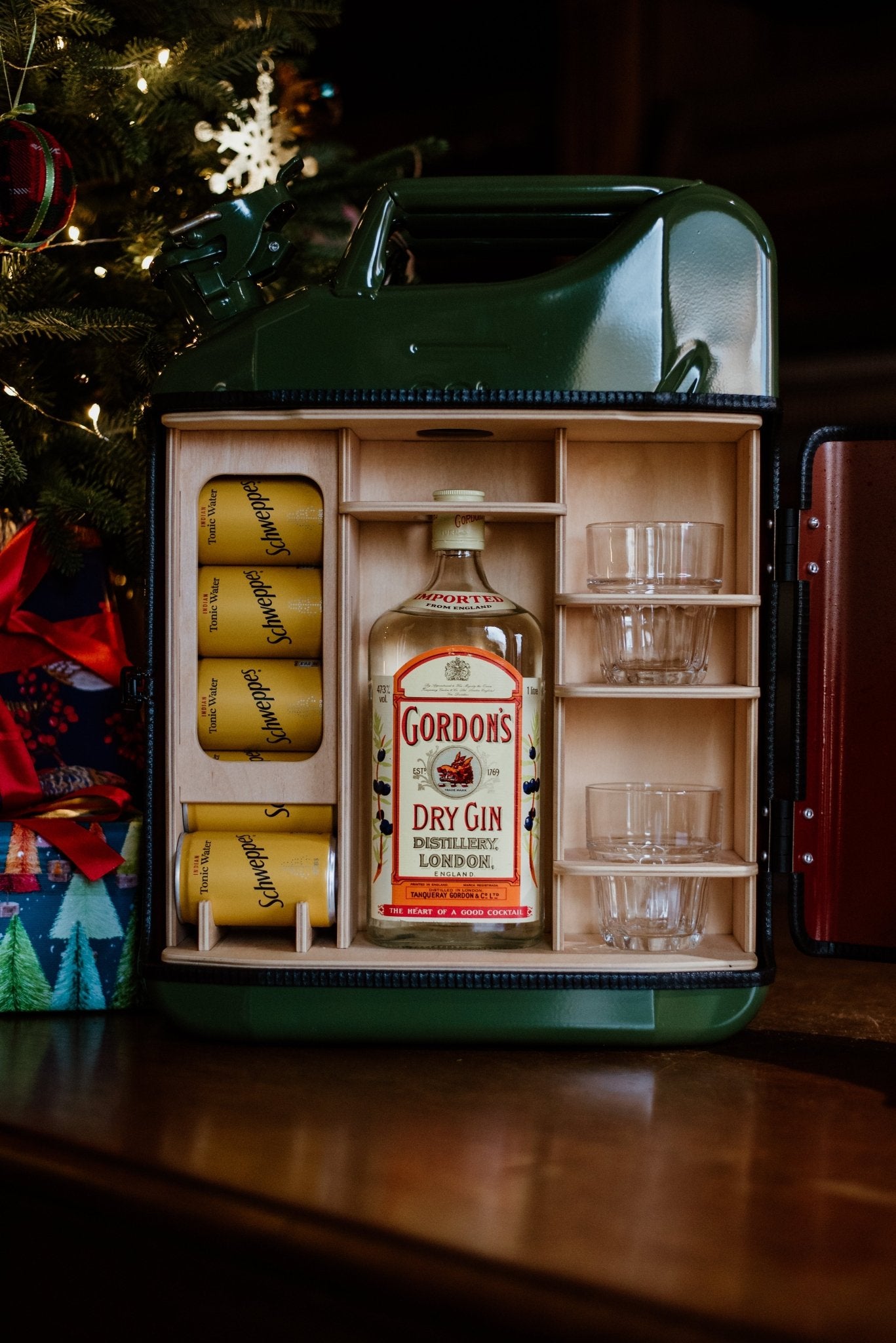 The Original Upcycled Mini Bar by Campbells of Beauly is a handmade jerry can mini bar containing Gordons Dry Gin, four Schweppes tonic cans, and four glass tumblers, displayed beside a decorated Christmas tree and gifts.
