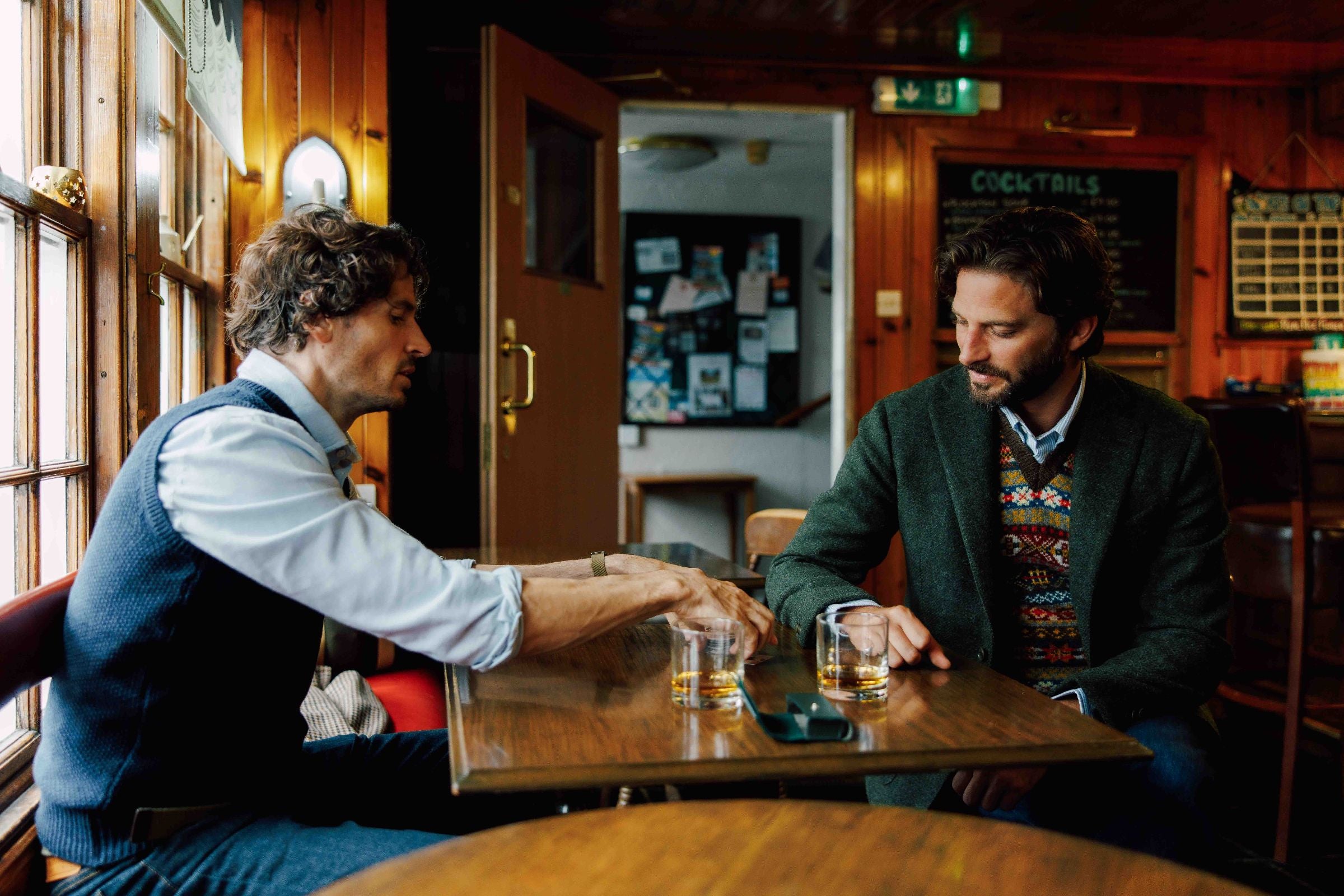 Two men sit at a wooden table in a cozy pub, each with a glass of whiskey. One gestures while talking; the other listens. Warm light streams through a window, and a cocktail menu is visible on the wall behind them.