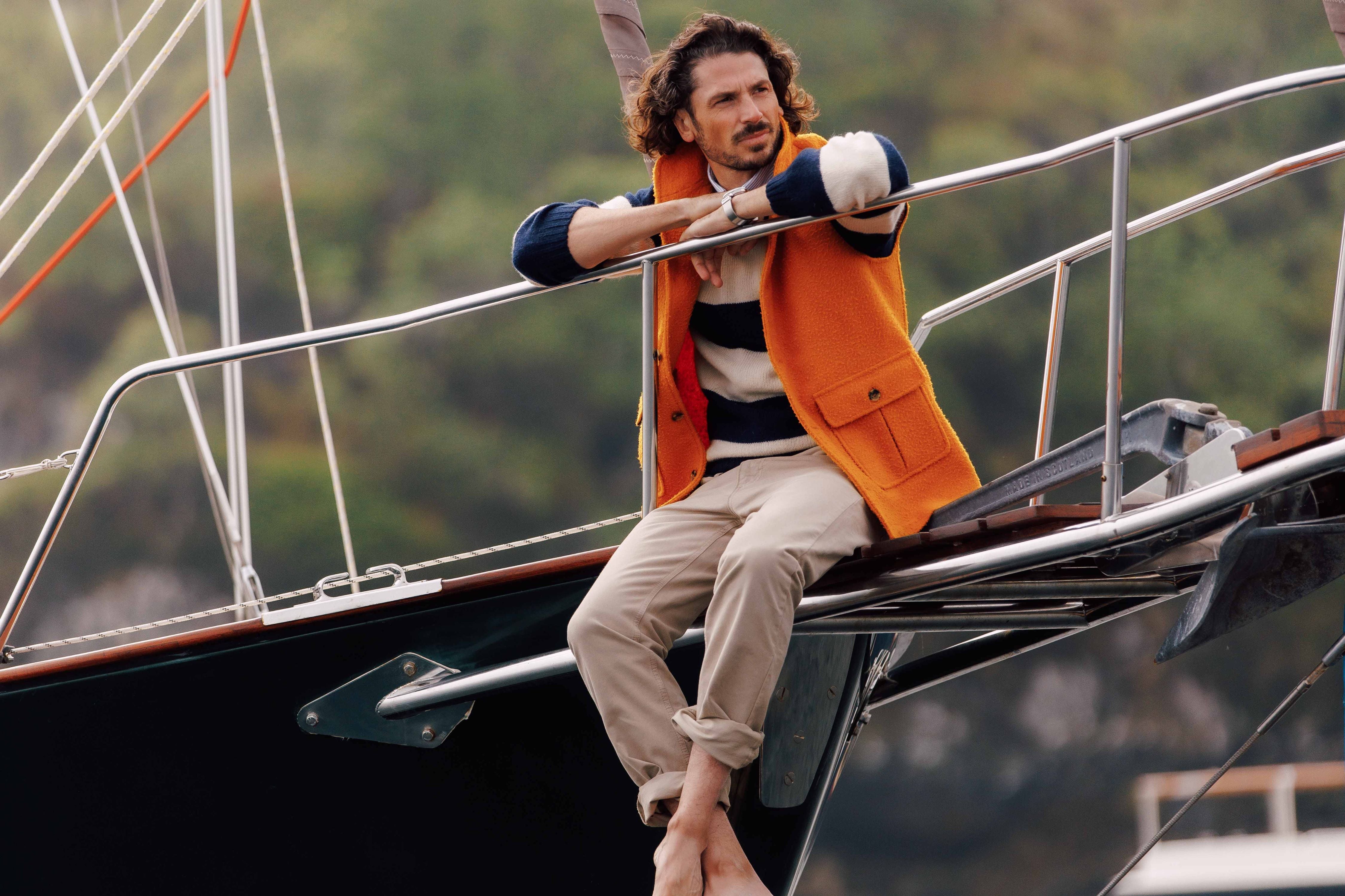 A man with curly hair, wearing an orange vest and striped sweater, sits barefoot on the bow of a sailboat, leaning on the railing with greenery blurred in the background.
