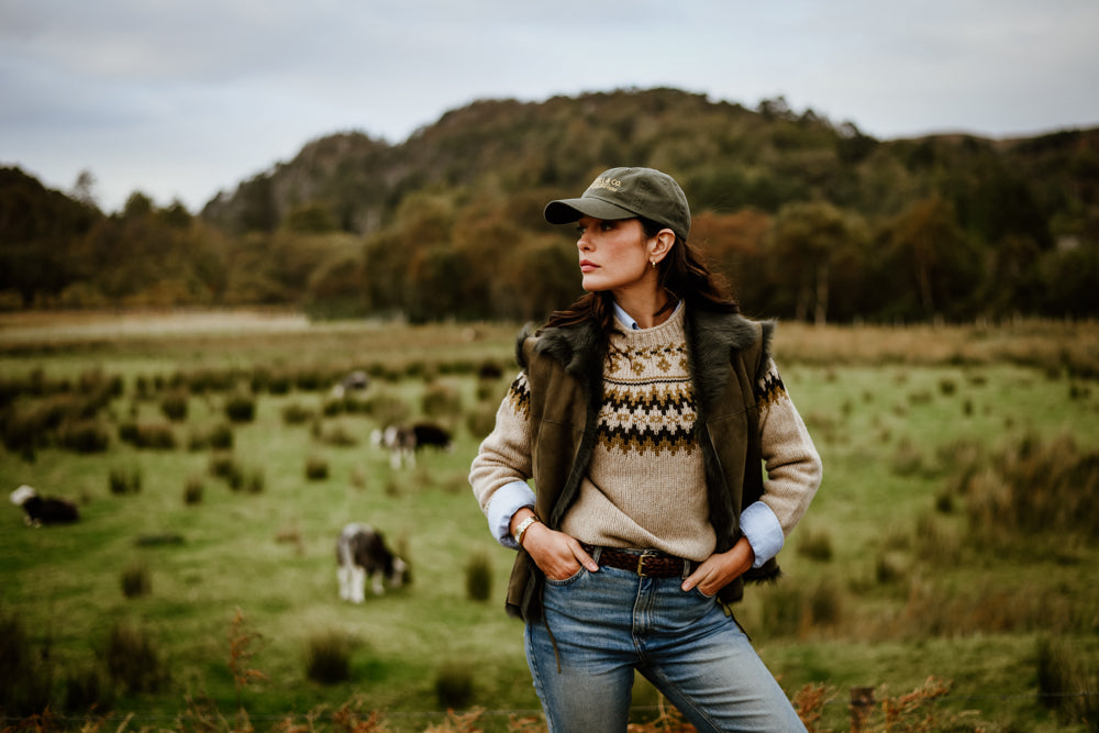 A woman in a cap, sweater, and jeans stands in a grassy field with cows grazing in the background and wooded hills under a cloudy sky.