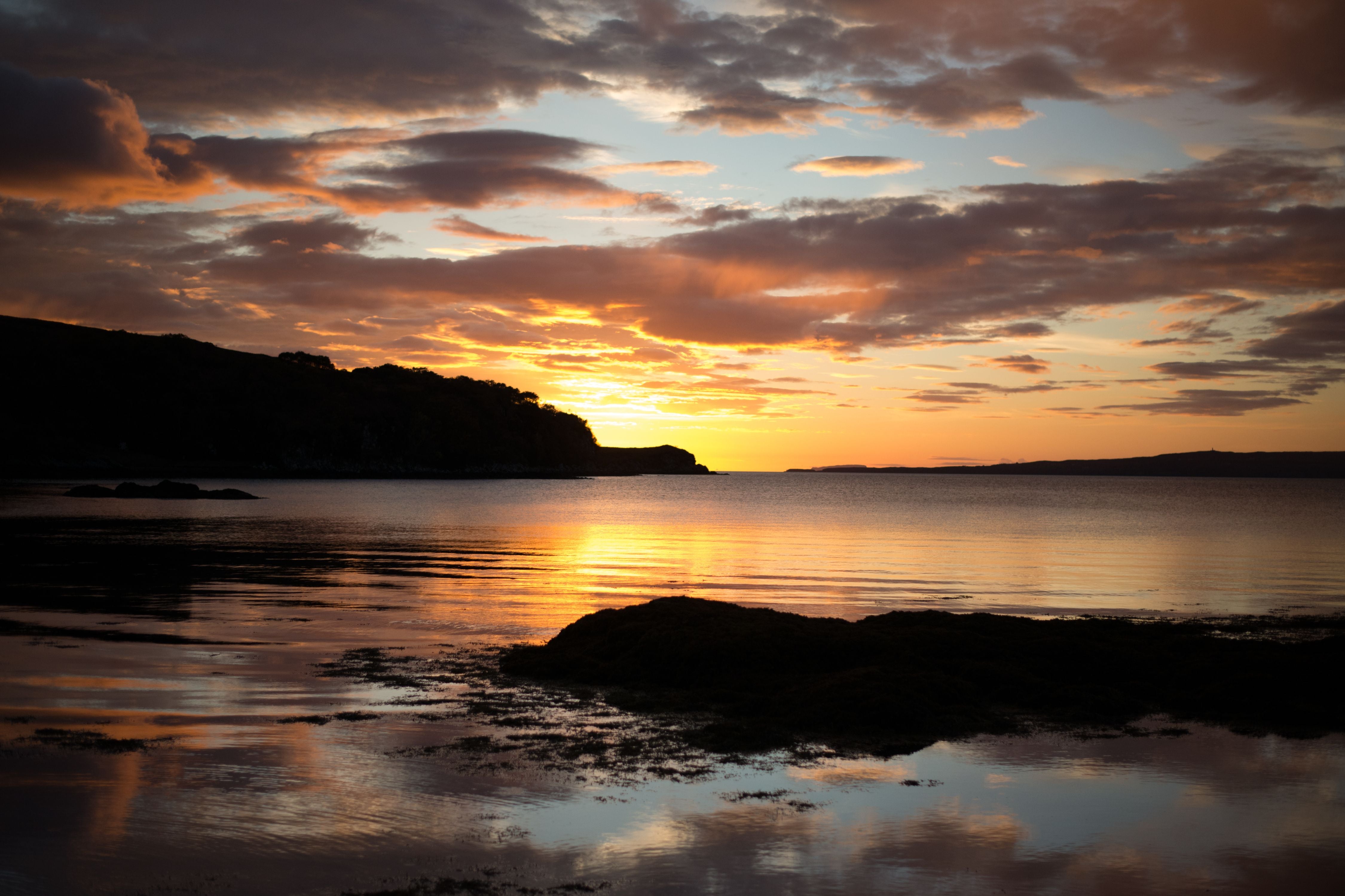 A serene sunset over a calm lake, with orange and yellow light reflecting on the water. Silhouetted hills and scattered clouds add depth to the peaceful landscape.