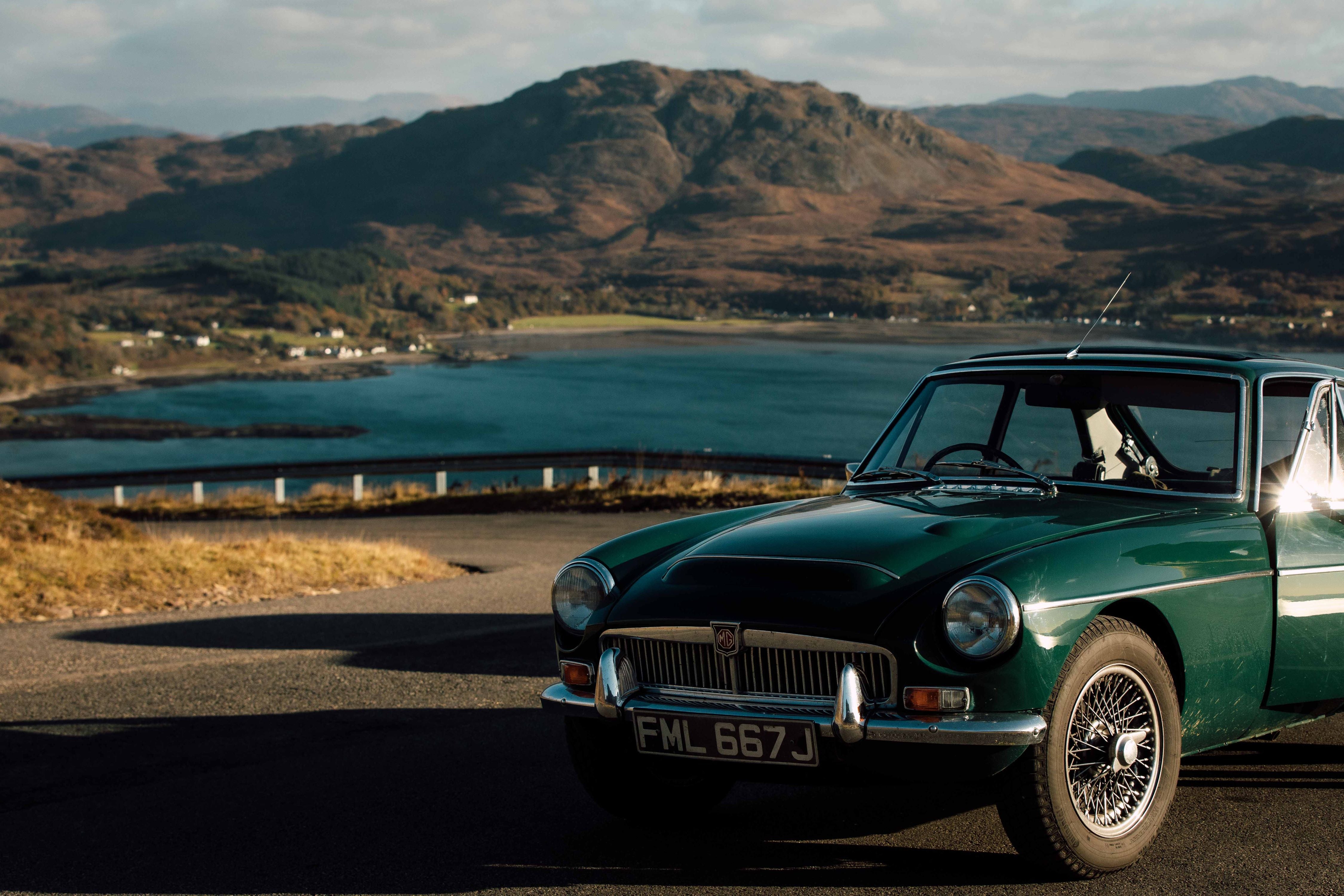 A classic green MG car parked on a scenic road overlooking a blue lake with rugged mountains in the background under a partly cloudy sky.