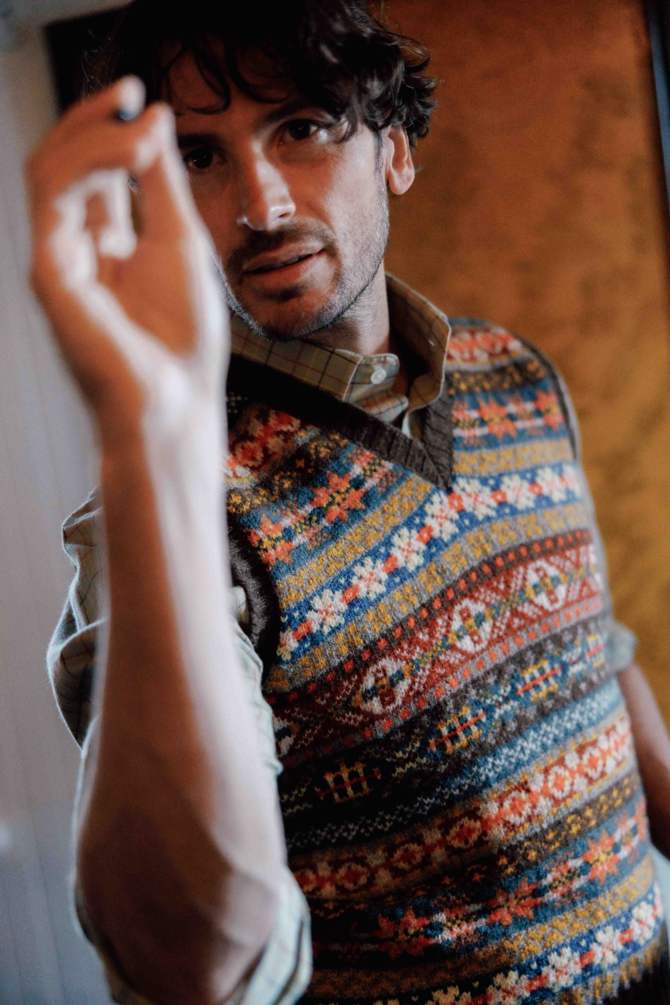 A man wearing a colorful patterned sweater vest and plaid shirt stands indoors, holding his arm up and appearing to write or draw on a surface in front of him, looking intently at the camera.