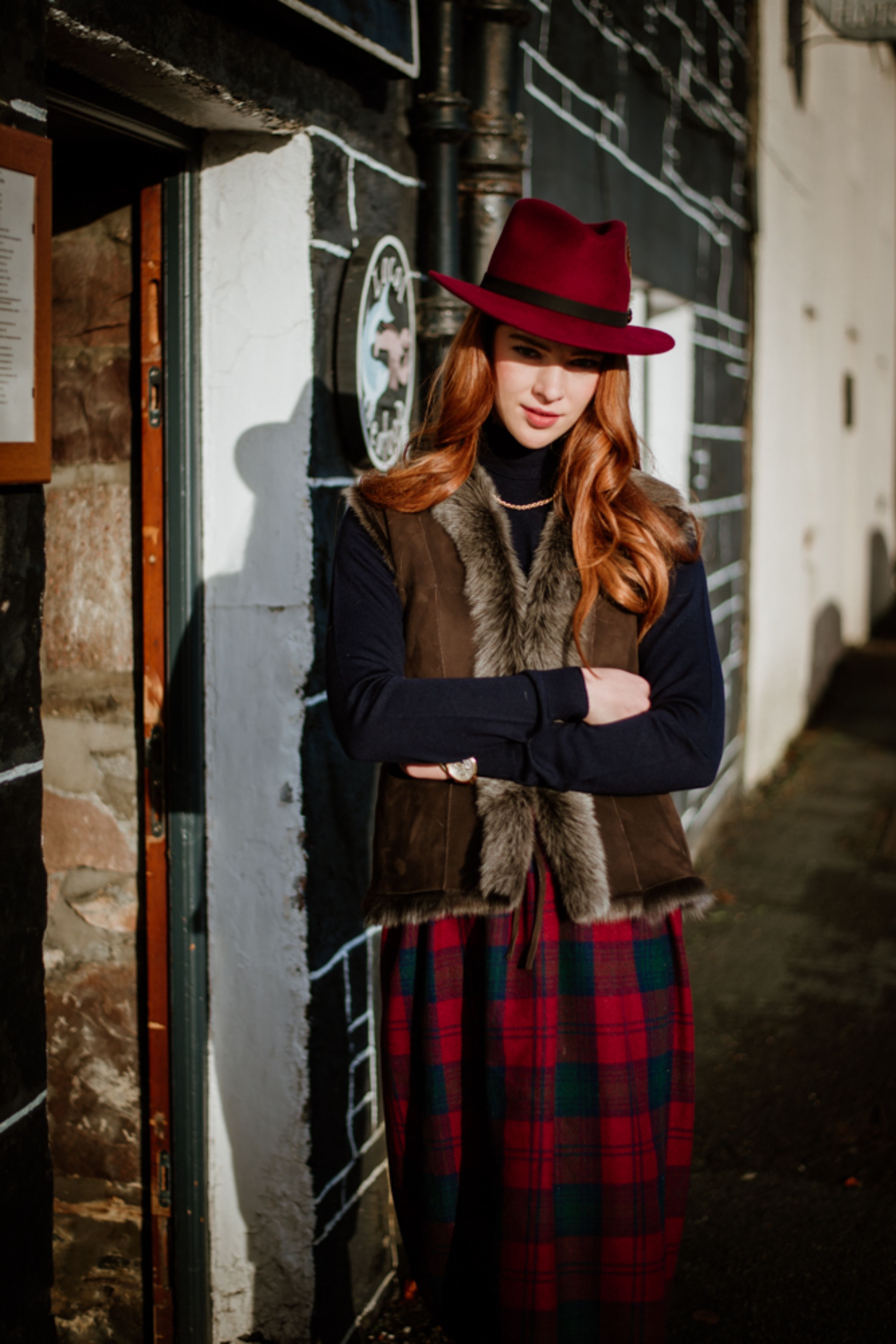 A woman with long red hair wearing a red hat, fur-trimmed vest, black turtleneck, and red plaid skirt stands with arms crossed outside by a black and white wall, partially in sunlight.