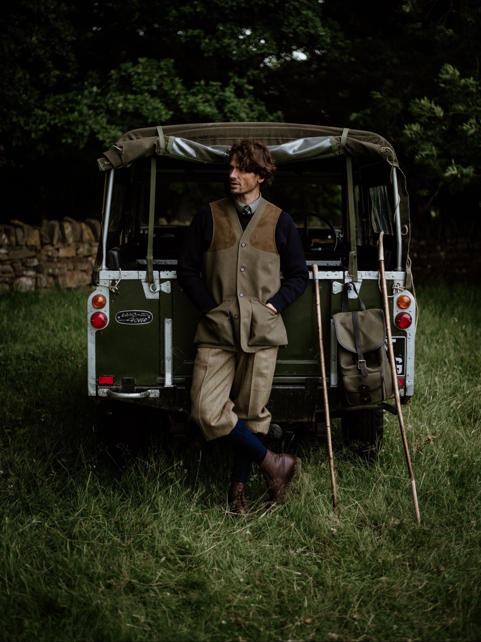 Dressed in vintage country style and a Campbell’s of Beauly Cotton Shooting Waistcoat, a person leans against a green Land Rover surrounded by walking sticks, a satchel, grassy terrain, dense foliage, and an old stone wall.