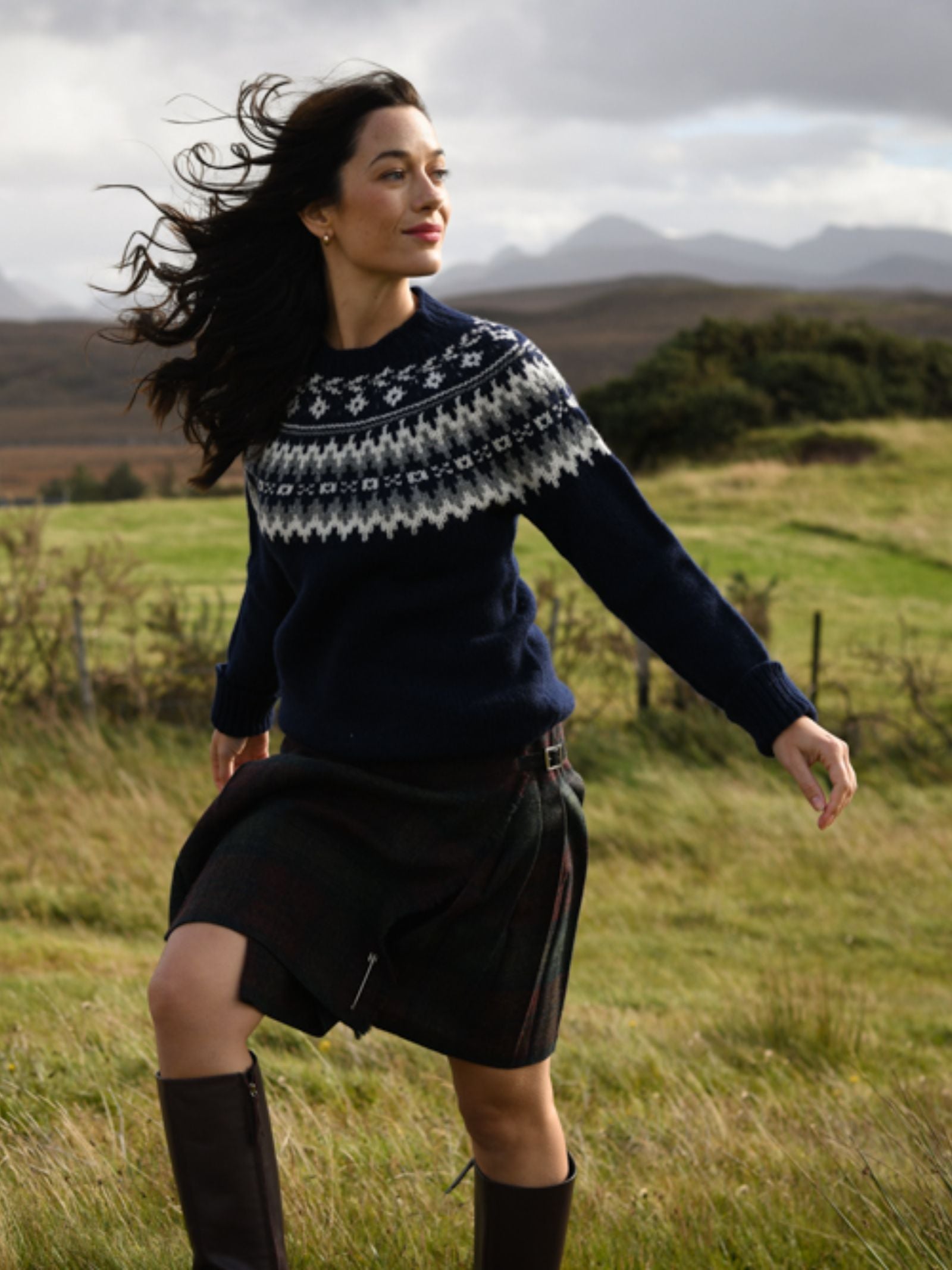 A woman with long, dark hair, wearing Campbell's of Beauly Auld Stock Chunky Fairisle Jumper, a plaid skirt, and brown boots walks through a grassy field with hills and mountains behind her under a cloudy sky.