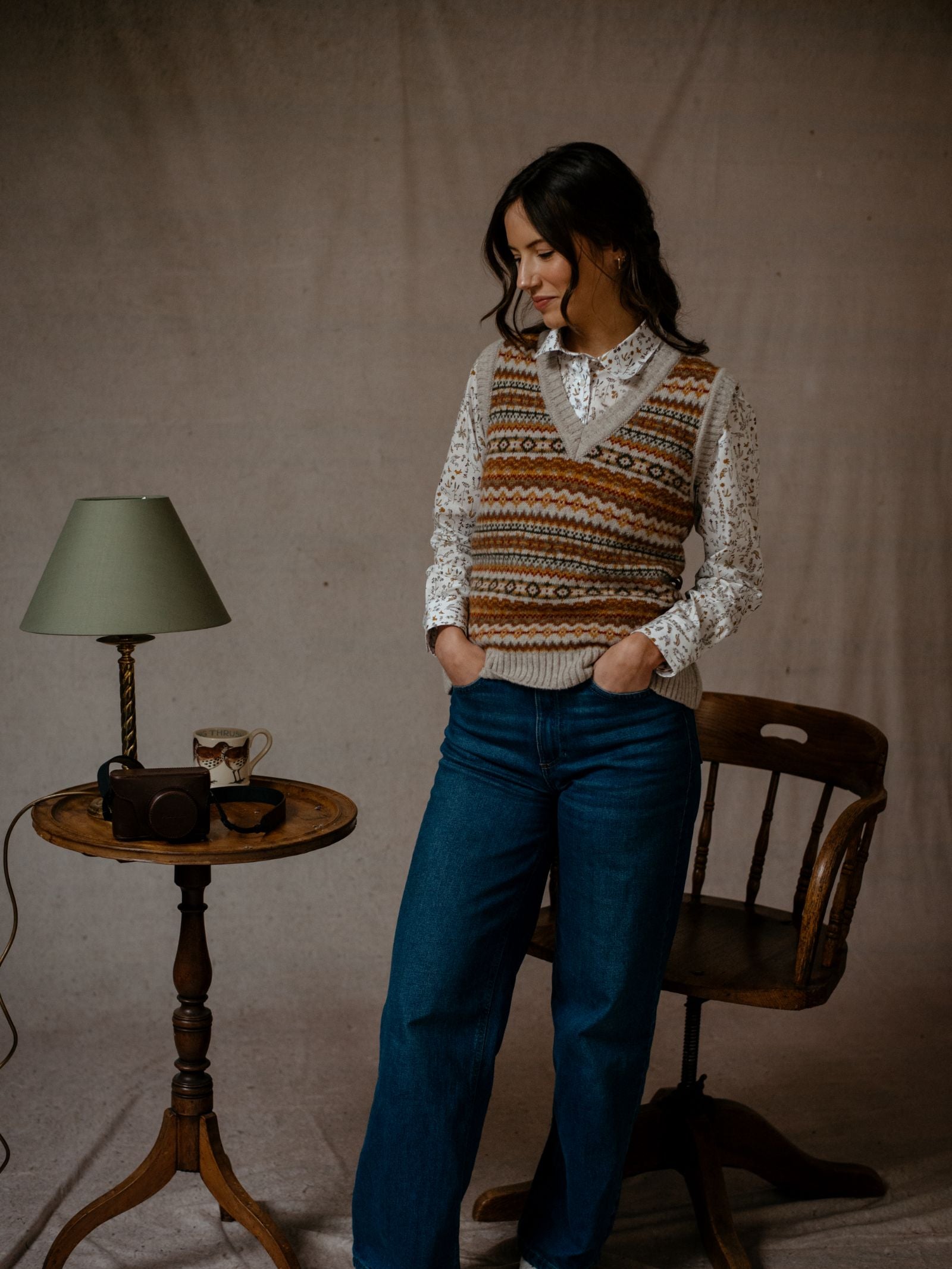 A woman indoors stands by a round wooden table with a green lampshade, mug, and camera. She's wearing Campbell's of Beauly's Auld Stock Fairisle V-Neck Tank over a white blouse and blue jeans, hands in her pockets. A wooden chair is nearby.