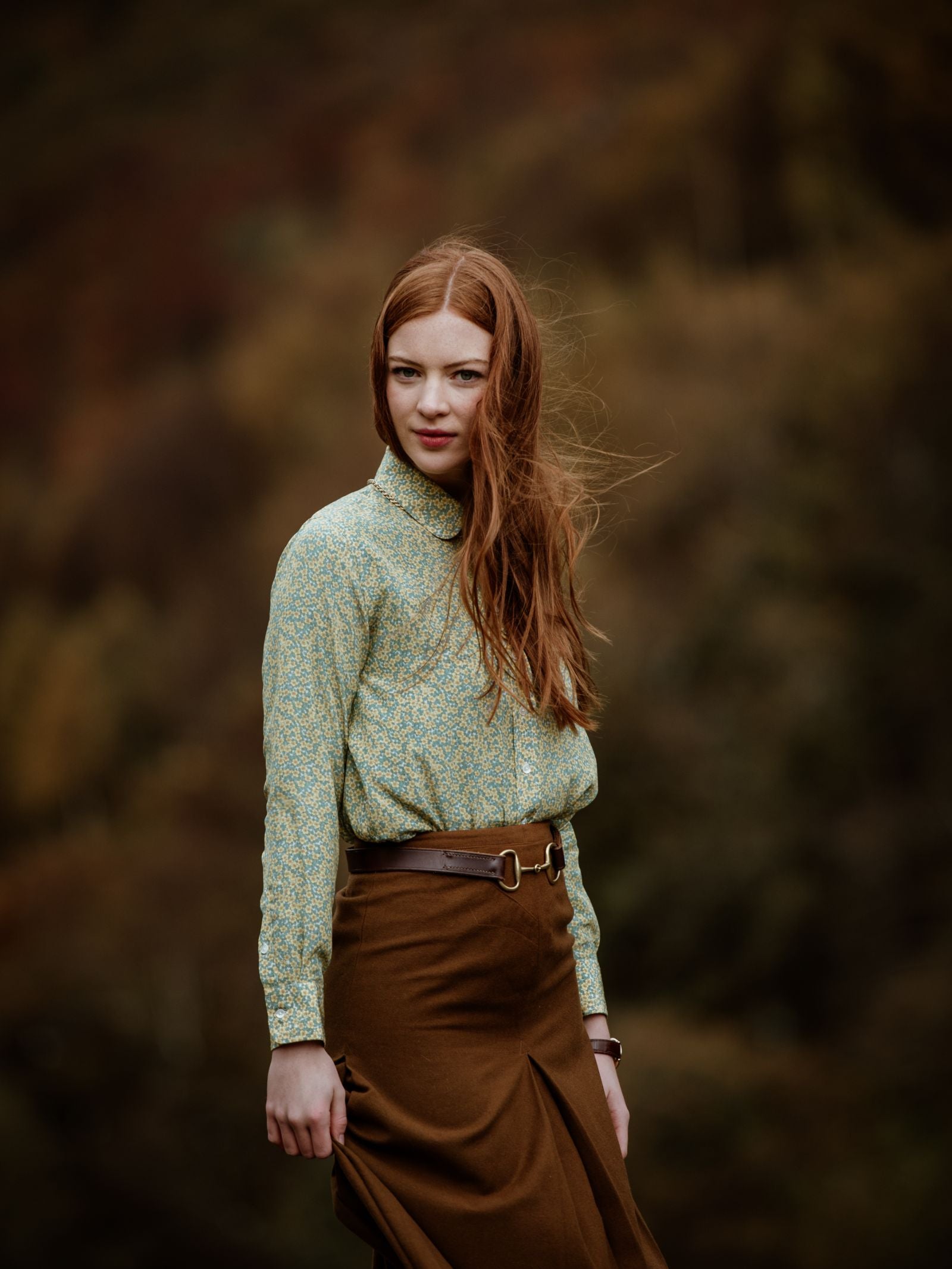 A young woman with long red hair stands outdoors in a Campbell's of Beauly Auld Stock Liberty Print Silk Shirt and a belted brown skirt. The blurred, earthy-toned background hints at an autumnal, natural setting.