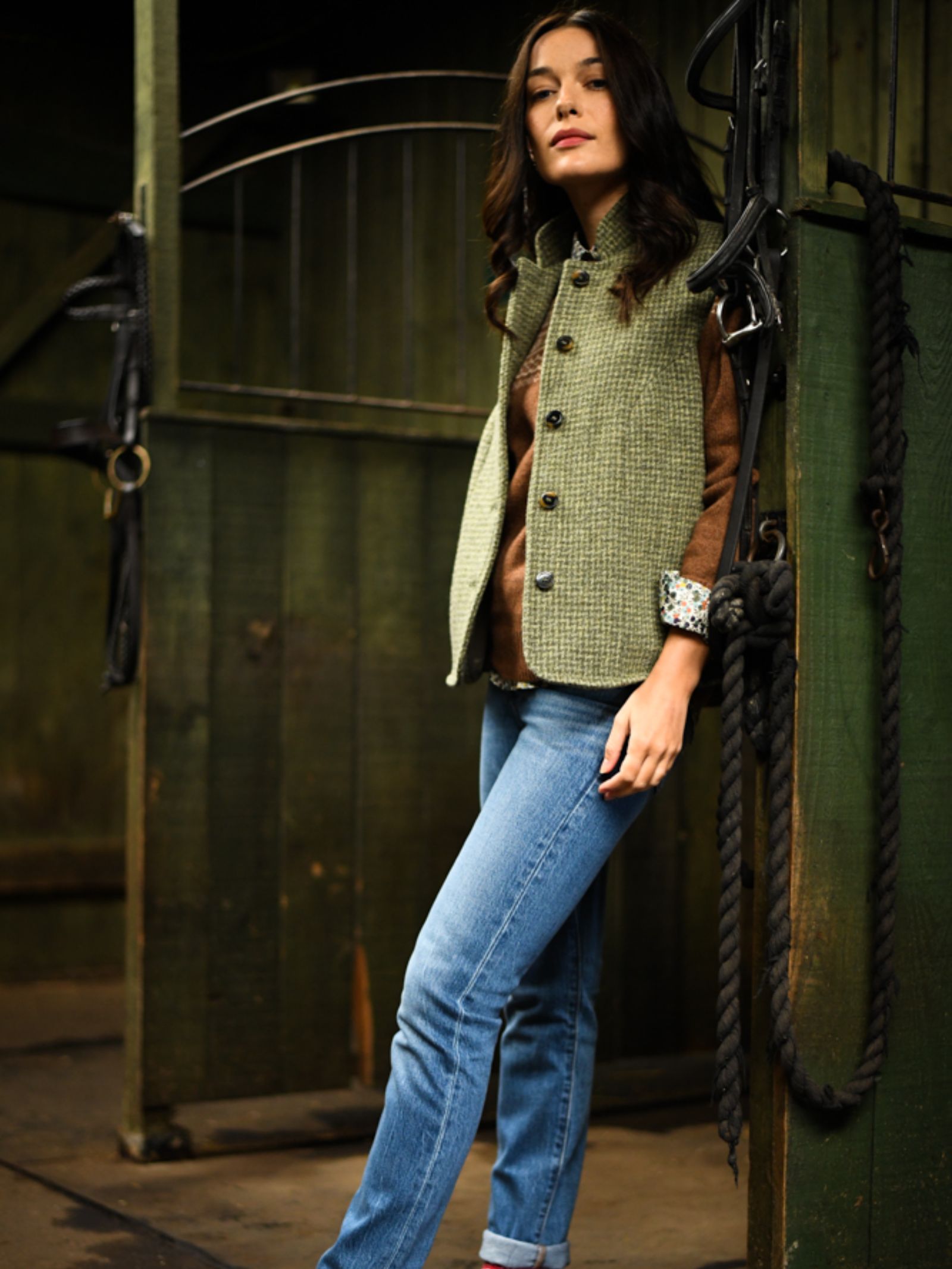A woman wearing the Campbell's of Beauly Auld Stock Harris Tweed Houndstooth Gilet, a brown sweater, and blue jeans stands in a rustic stable doorway with green wooden doors and hanging black ropes, resting one arm on the frame.