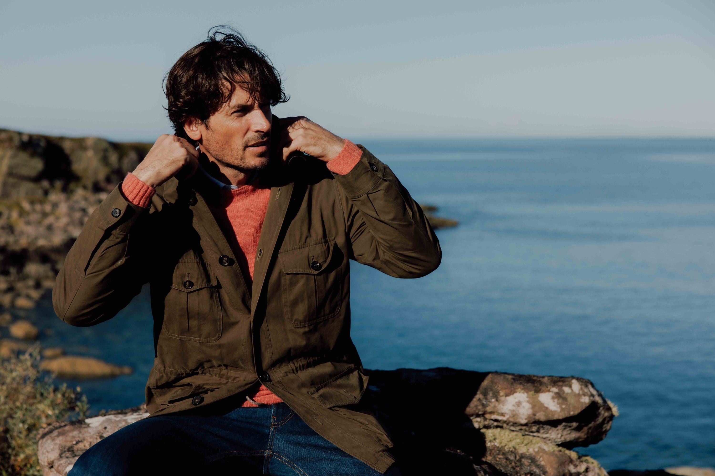 A man sits on rocks by the sea, adjusting the collar of his olive green jacket. He wears a red sweater underneath and blue jeans, with a calm water and rocky shoreline in the background on a clear day.