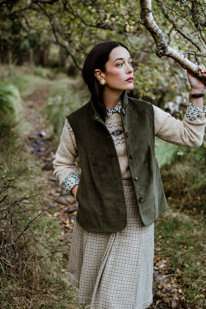 A woman in vintage-style clothing stands on a wooded path, holding a tree branch. She wears a green vest over a patterned shirt and a plaid skirt, with her hair down and red lipstick. The background is lush and leafy.