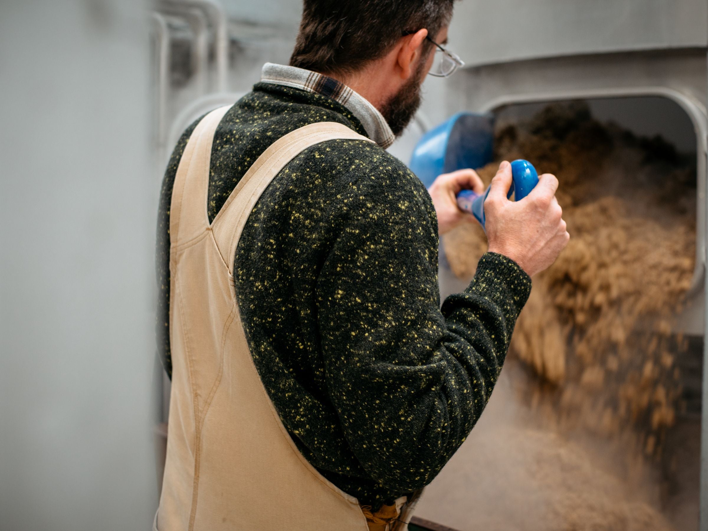 A person wearing a dark sweater and overalls pours a scoop of grains or feed into a large industrial machine or container, seen from behind in an indoor setting.