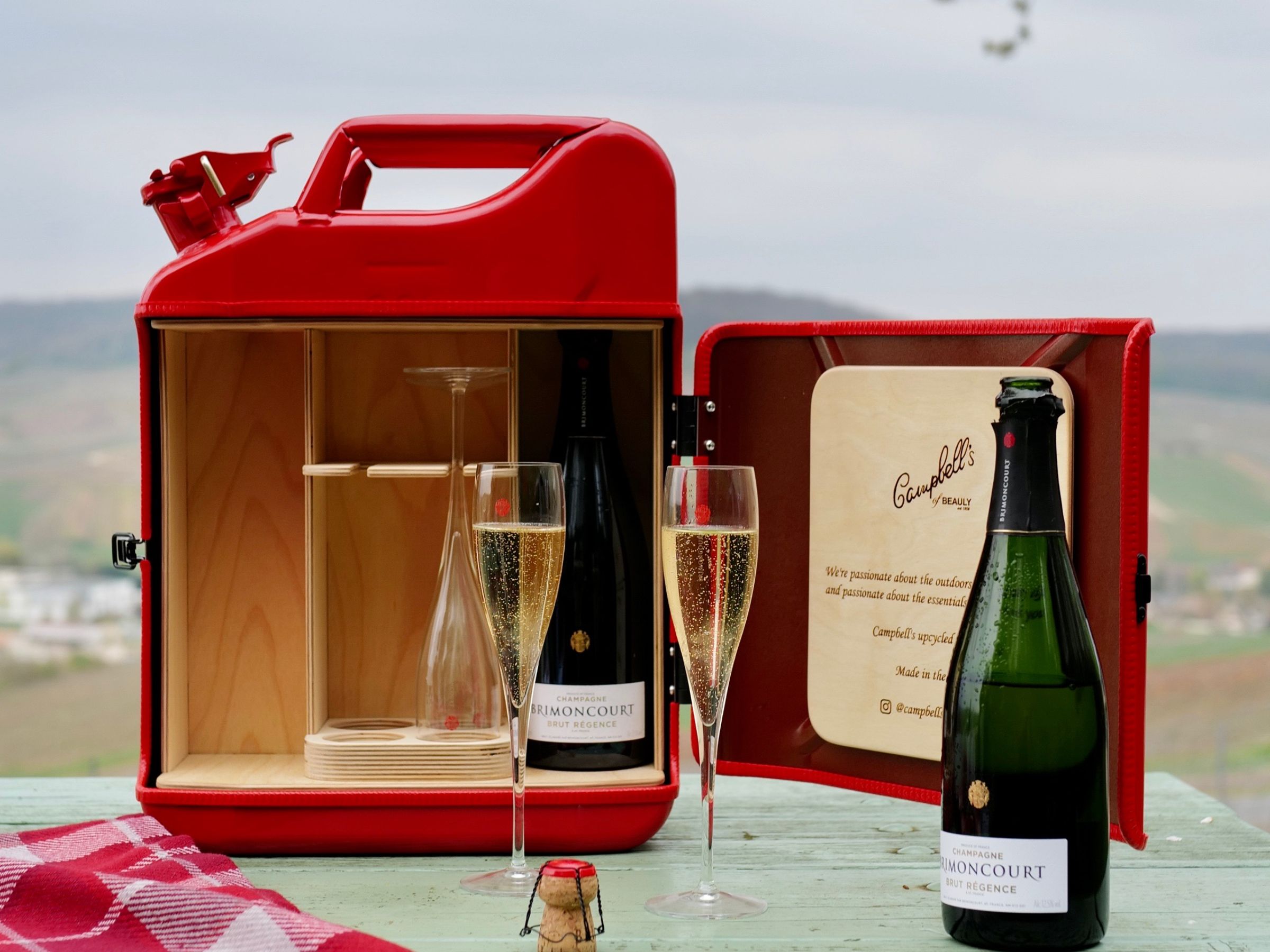 A red box styled like a fuel canister is open on a table, revealing two champagne glasses and a bottle inside. Nearby are another champagne bottle, two filled glasses, and a cork, with a scenic countryside in the background.