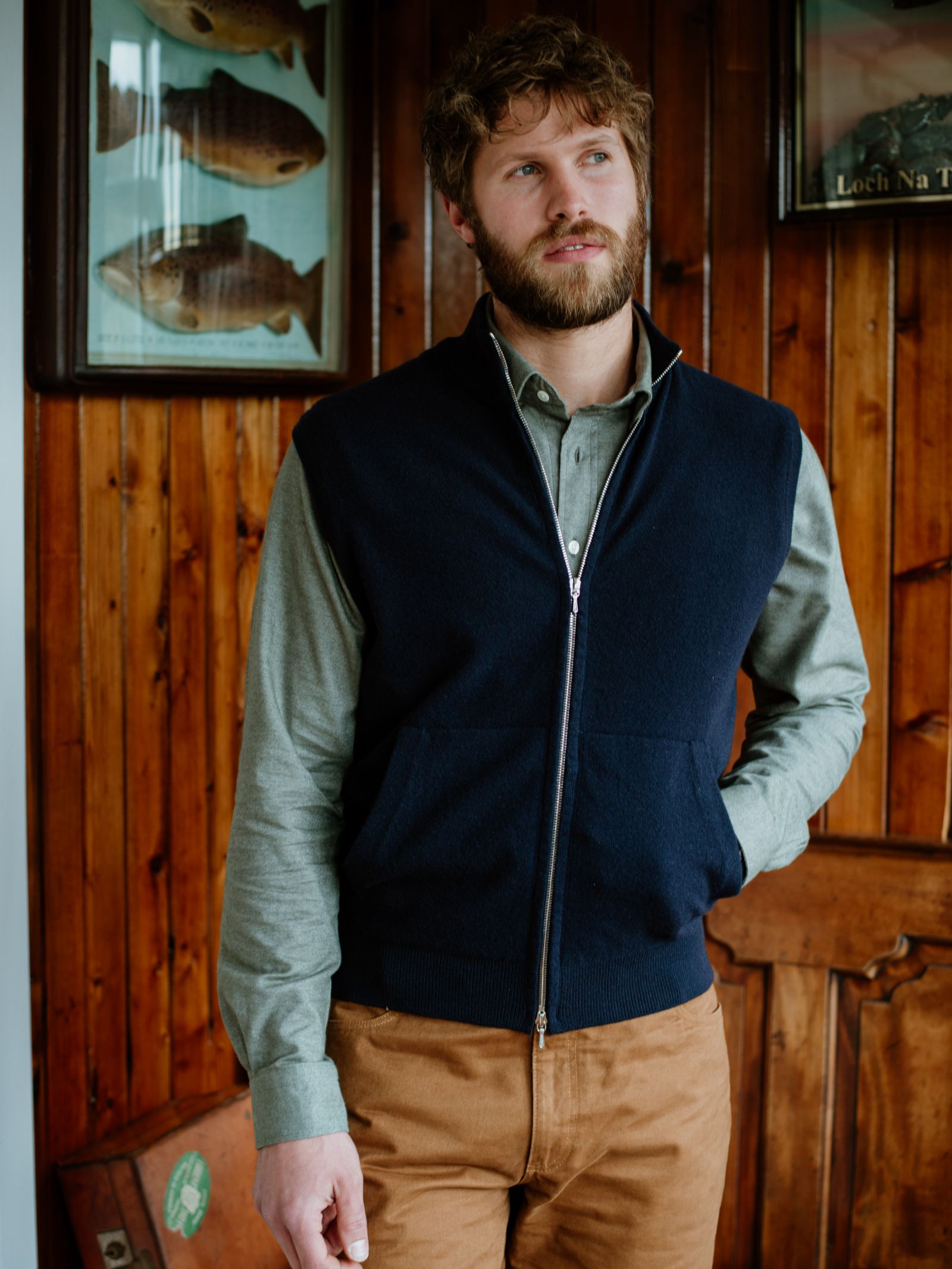 A bearded man stands indoors against a wooden wall, wearing a green button-up shirt, brown pants, and the Campbell's of Beauly Auld Stock Zip Gilet. Framed fish prints and a wooden door are seen in the background.