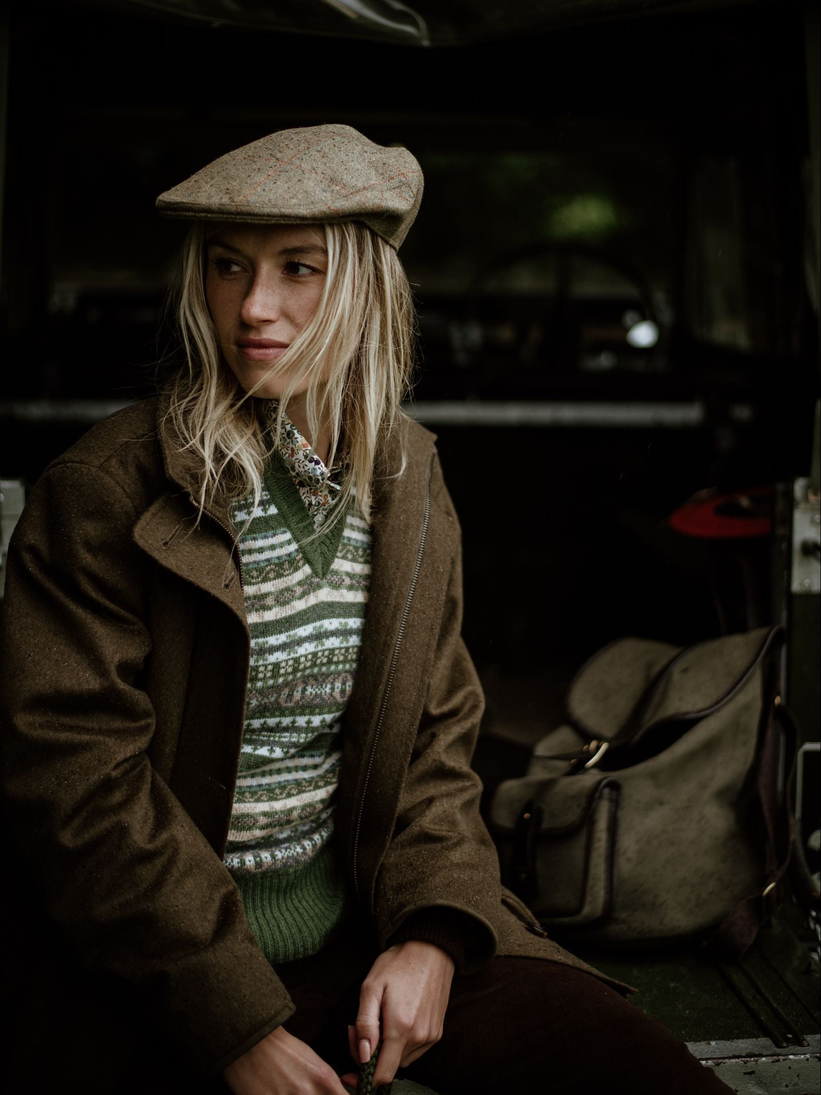 A blonde woman wears the Campbell's of Beauly Auld Stock Cashmere V-Neck Fairisle Slipover, a tweed cap, and a brown coat, sitting in a vehicle with a canvas bag by her side, gazing thoughtfully to the side.
