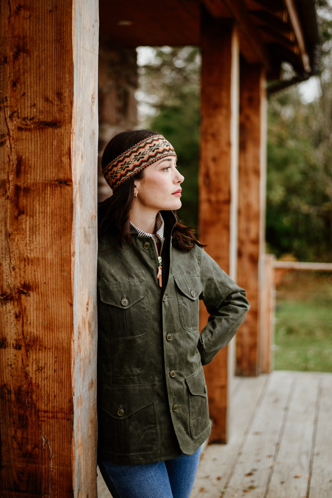 A woman wearing a green jacket, patterned headband, and jeans stands on a wooden porch, leaning against a post and looking thoughtfully into the distance. Trees and greenery are visible in the background.