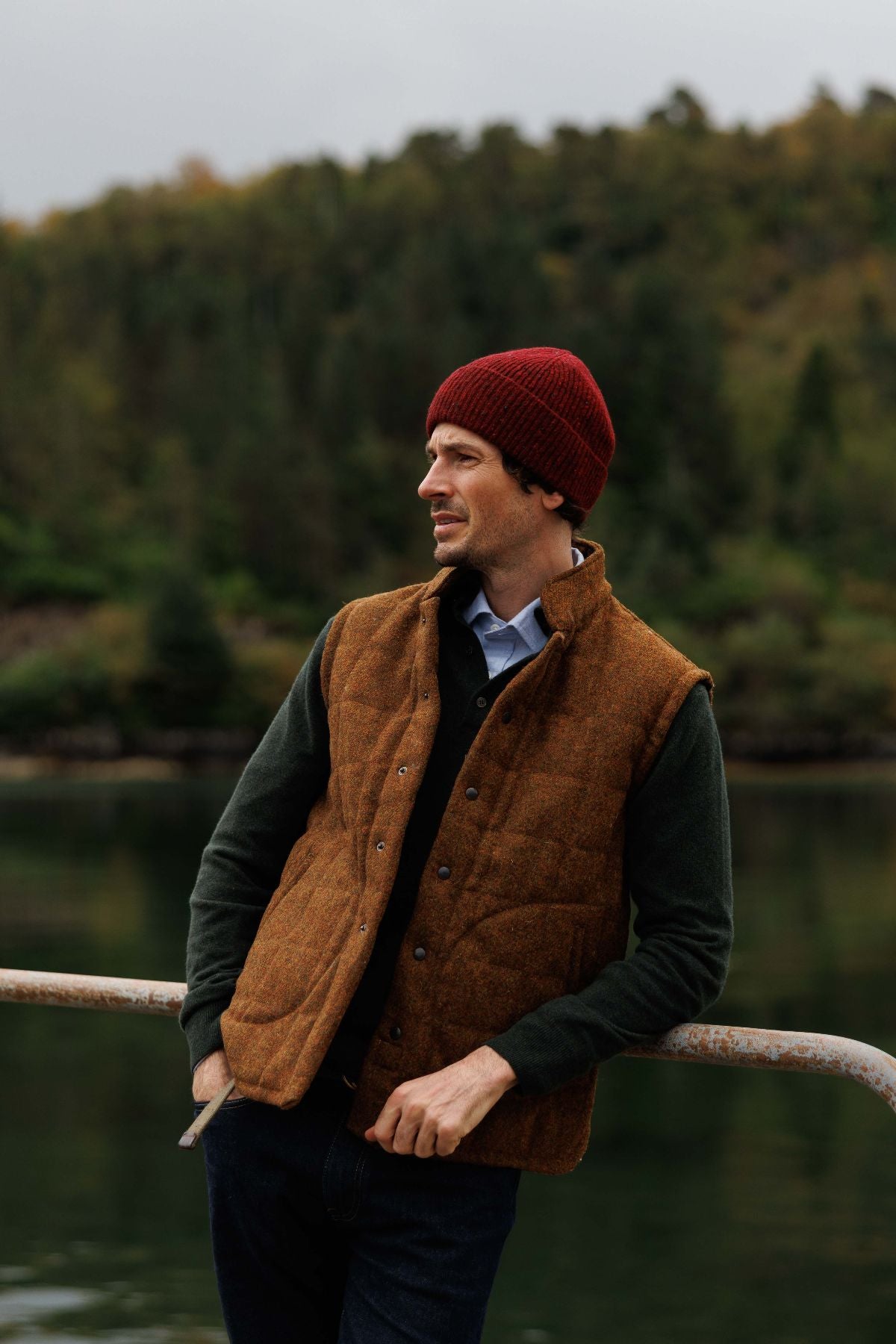 A man wearing a red knit beanie, a brown quilted vest, and a dark long-sleeve shirt stands outdoors by a metal railing with a lake and forested hills in the background.