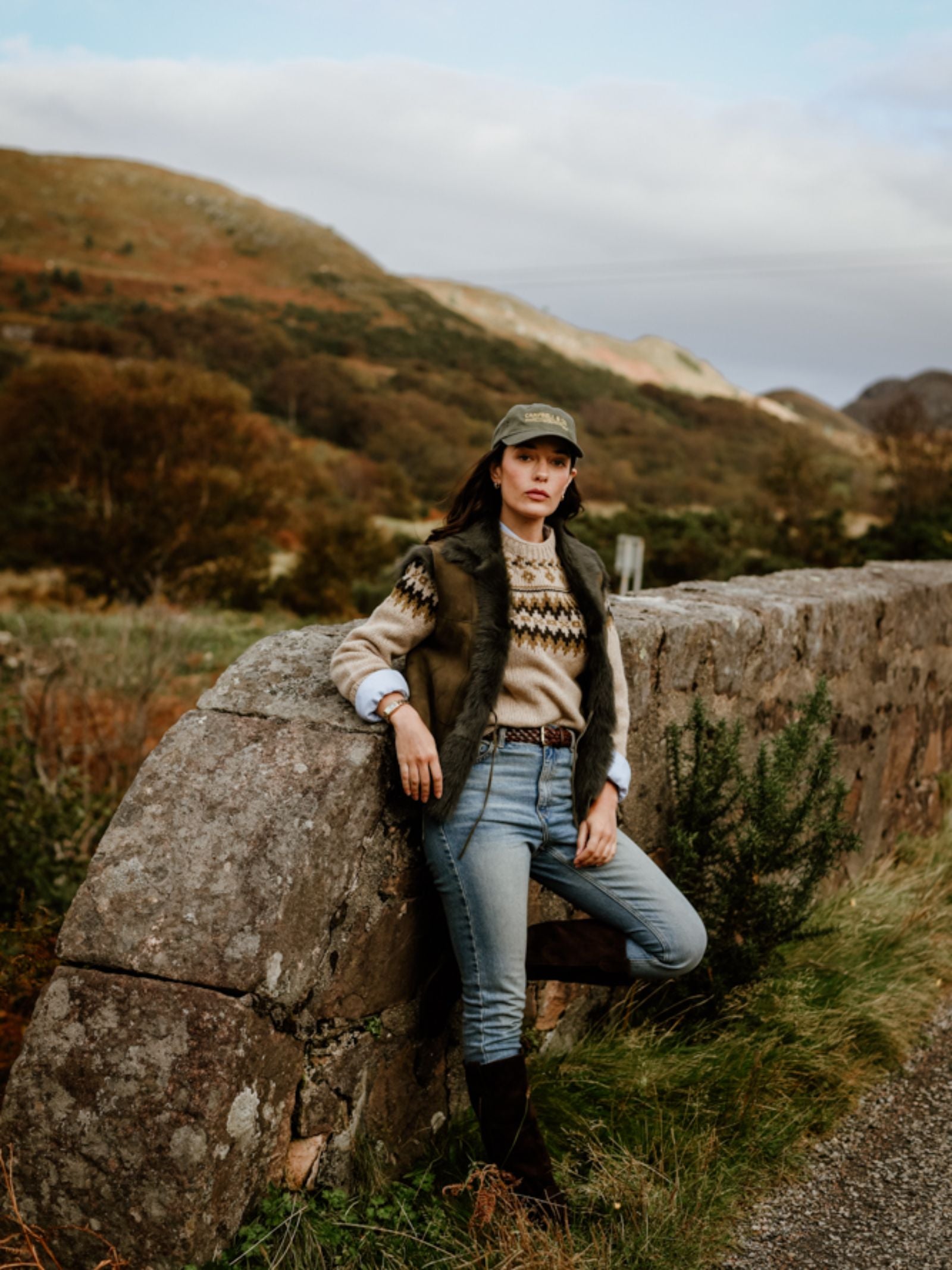 A woman in jeans, boots, a patterned sweater, and the Campbell's of Beauly Auld Stock Sheepskin Reversible Gilet leans against a stone wall with grassy hills and cloudy skies in the rural background.