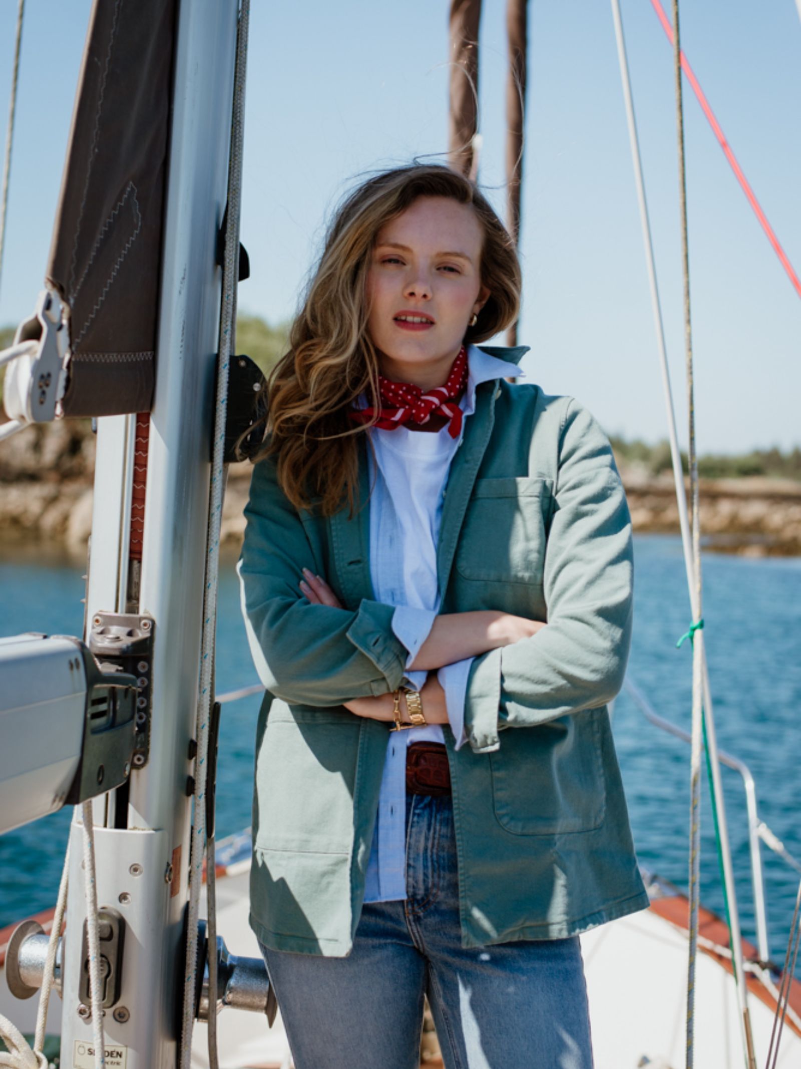 A woman with wavy blonde hair stands on a sailboat, wearing a light green jacket, white shirt, and a Campbells of Beauly Cotton Spotty Hank, its classic red design adding charm against the blue water and coastline.